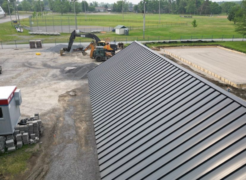 Gravel construction site with excavator beside a large gray metal roof and a concrete slab