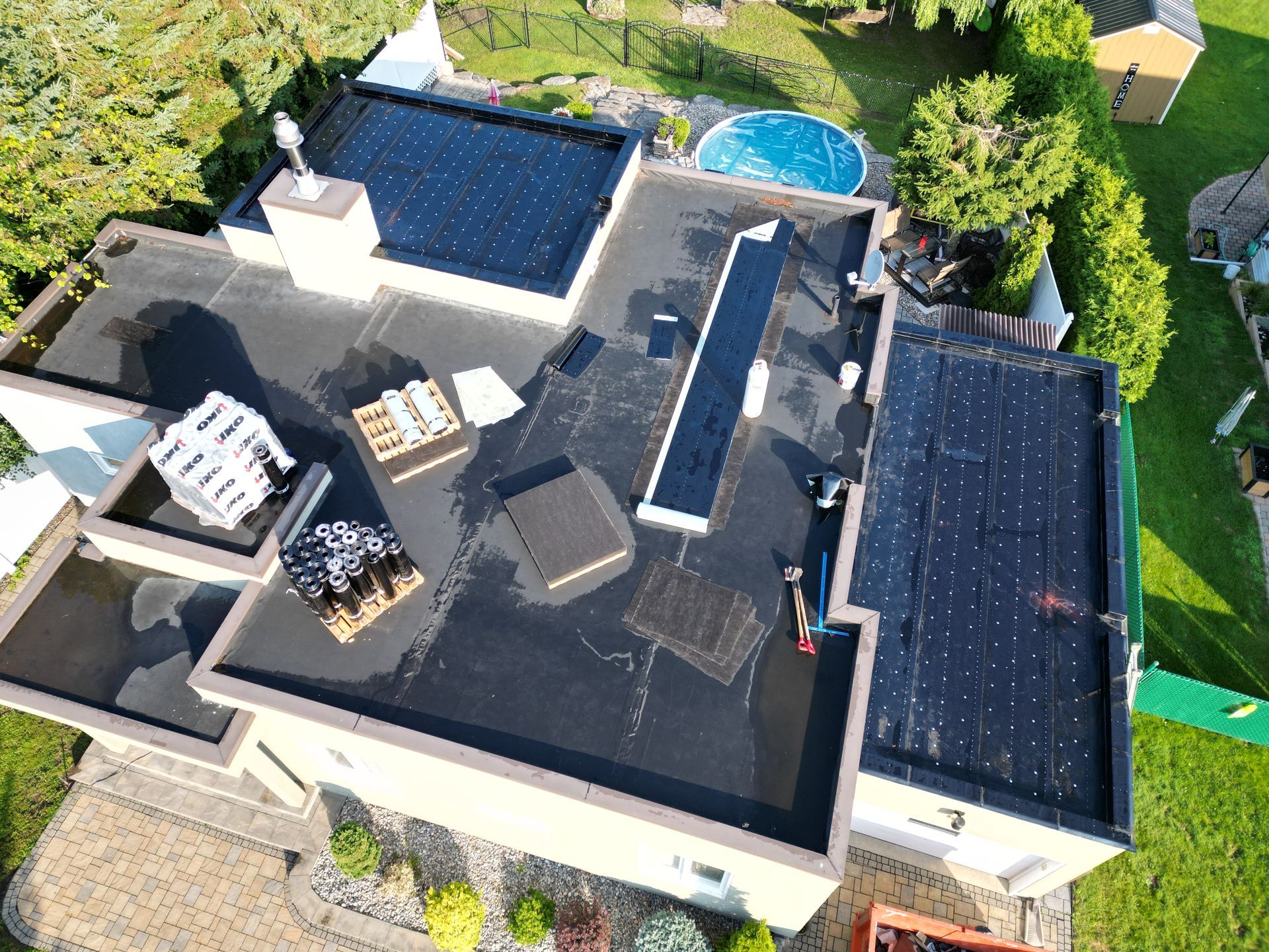 Aerial view of a house with a flat dark roof, patio furniture, a pool, and surrounding greenery