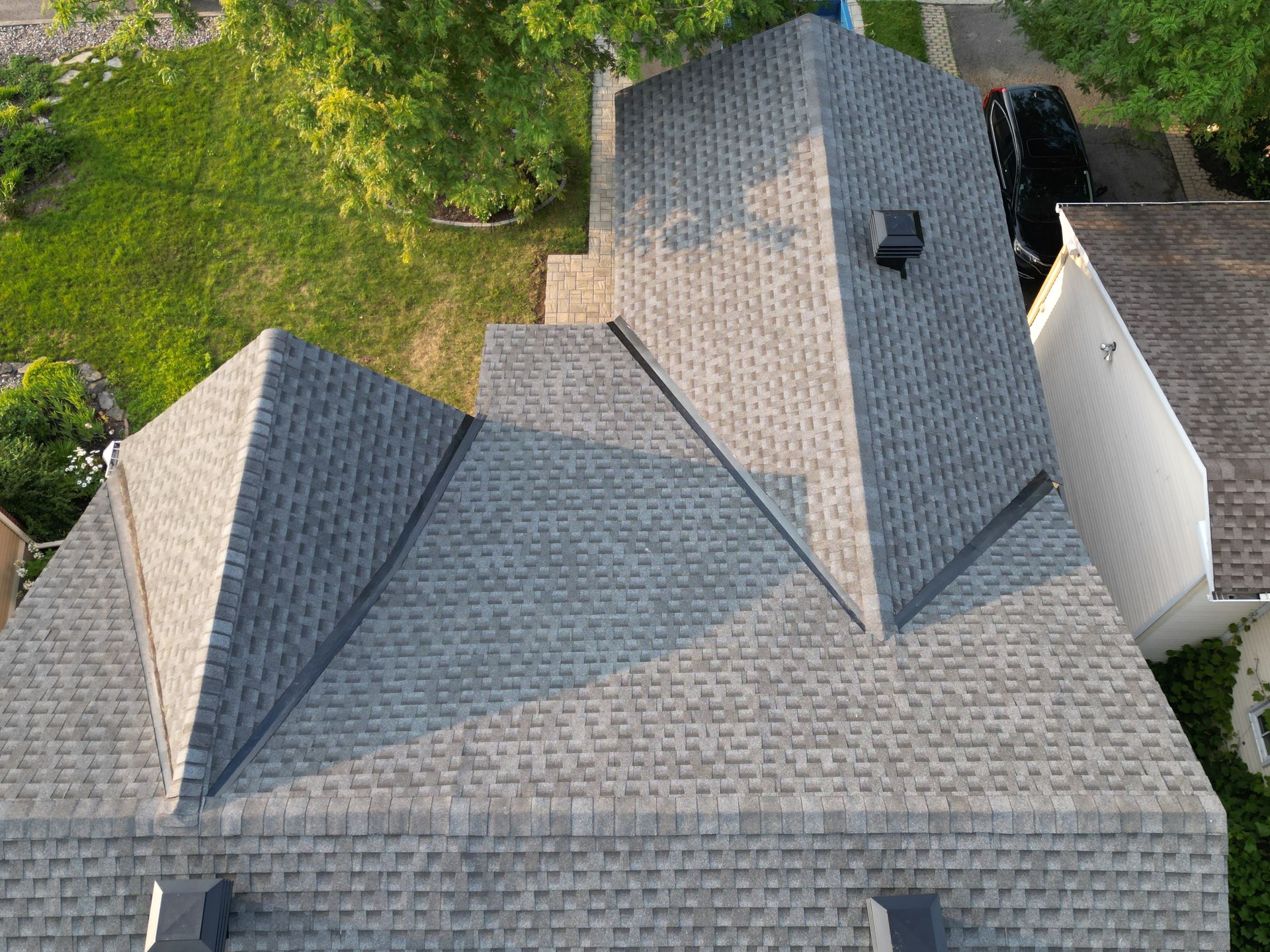 Aerial view of a gray shingled roof with multiple gables and a chimney, surrounded by trees.