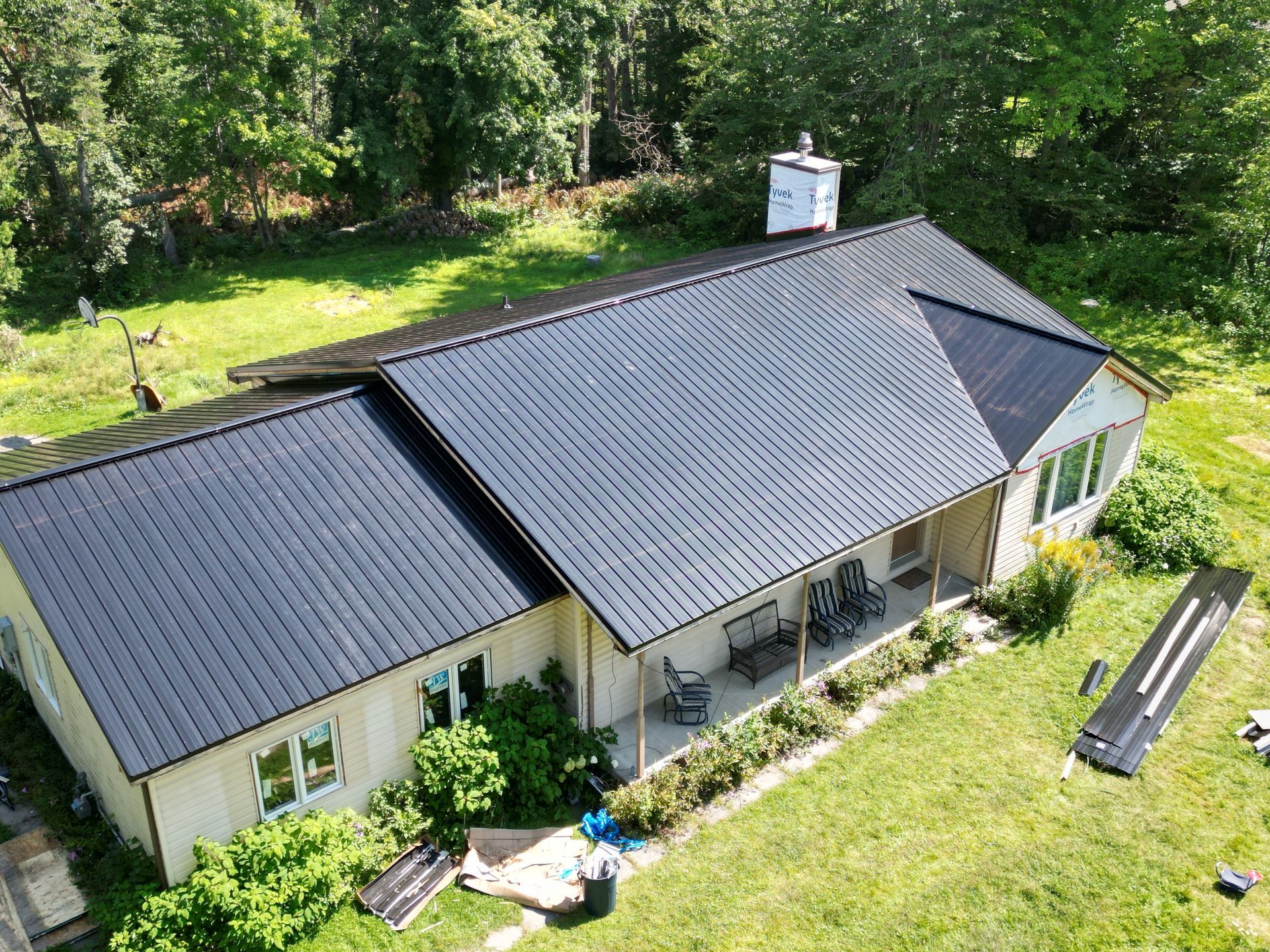 Aerial view of a house with a dark roof, white chimney, and green yard surrounded by trees.