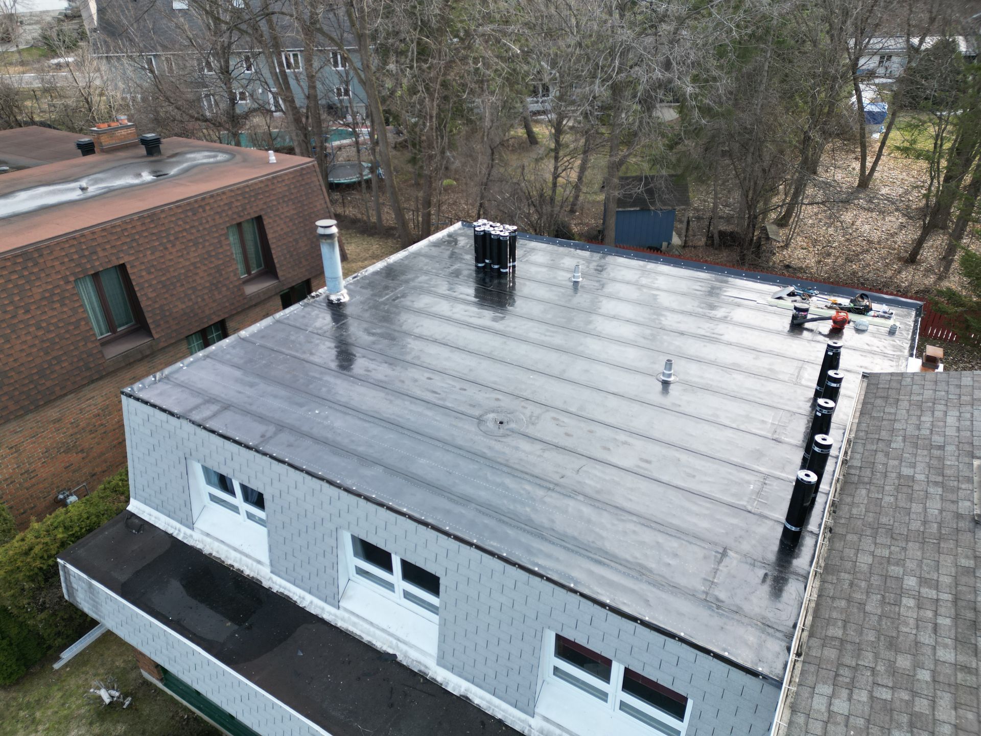 Aerial view of a flat-roofed building with vents, surrounded by trees and neighboring brick buildings.