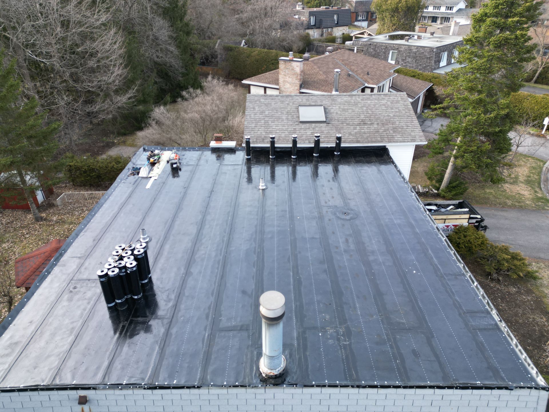 Flat black rooftop with vents and skylights, viewed from above in a wooded residential area