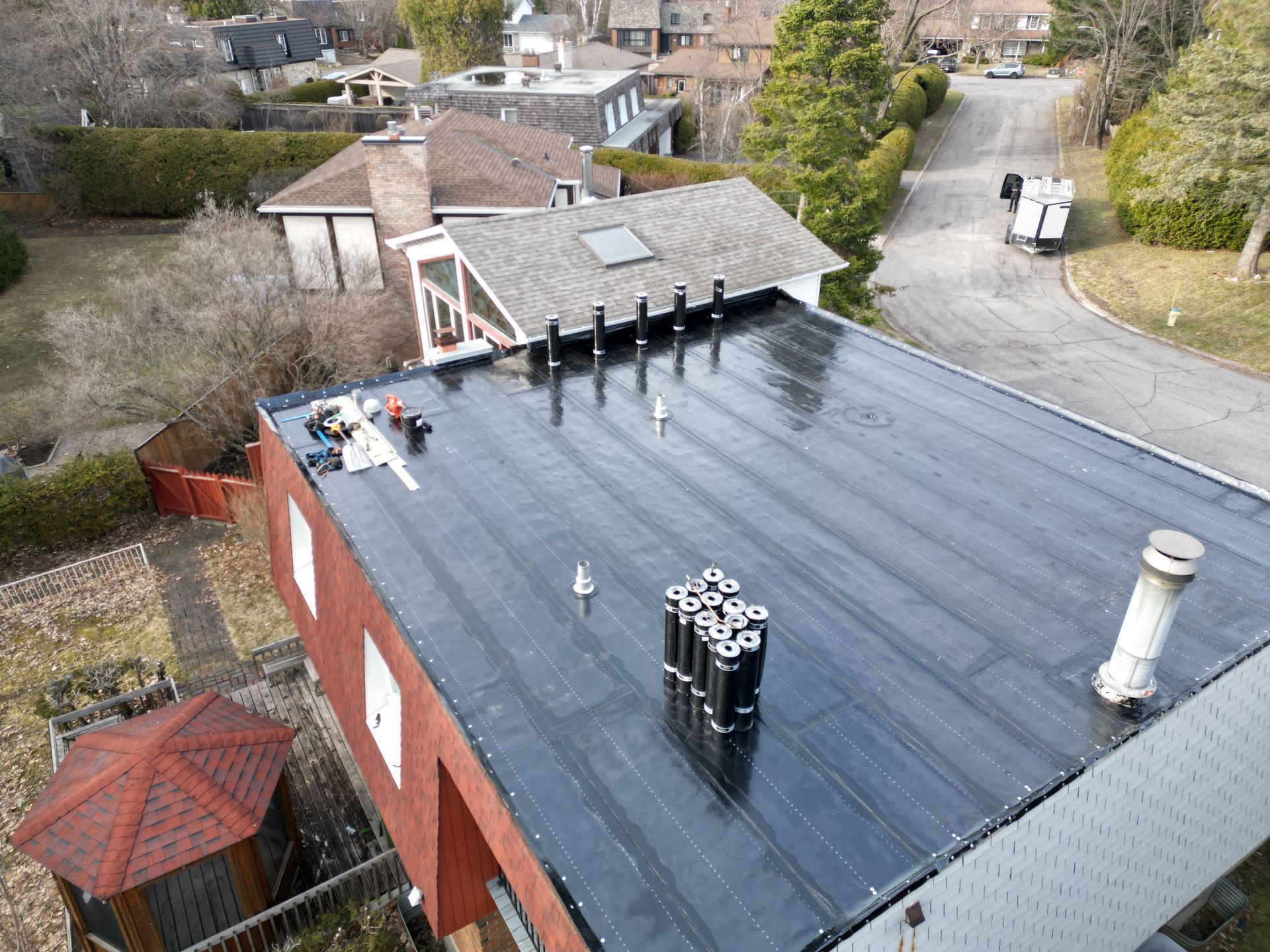 Aerial view of a red-brick building with a black roof, rooftop vents, and nearby houses and trees.