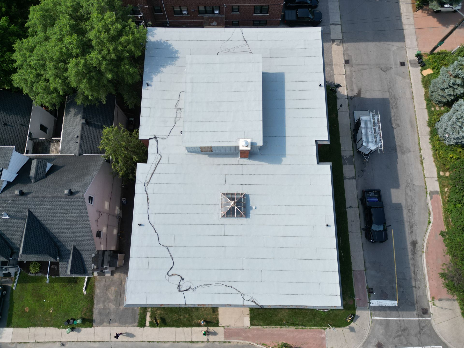 Aerial view of a flat-roofed building with neighboring houses, trees, and parked cars along a street.