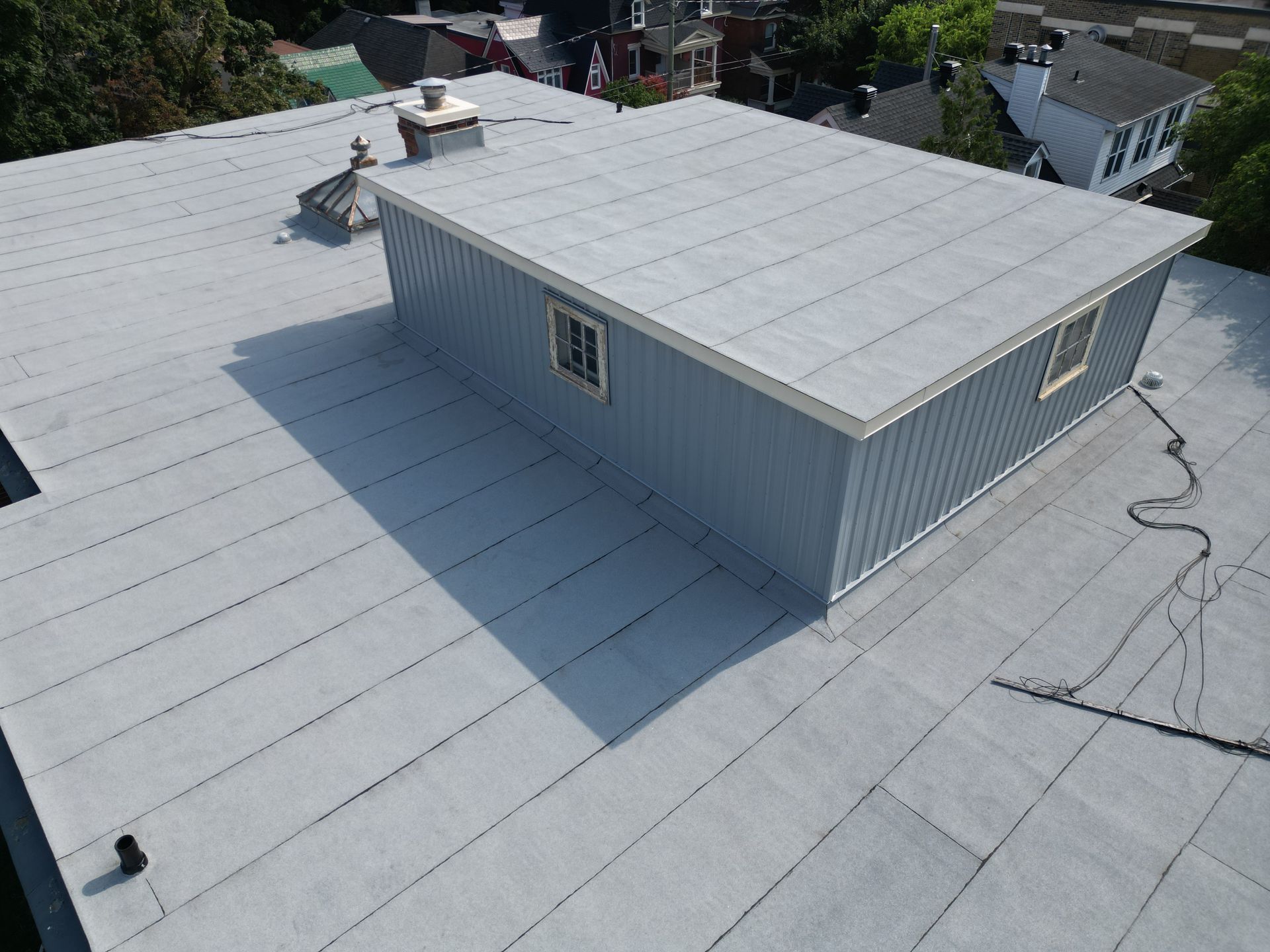Aerial view of a flat gray rooftop with a small rooftop structure and nearby residential houses.