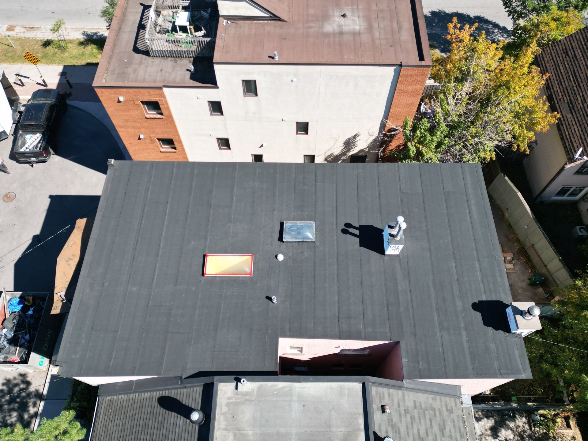 Aerial view of a flat black roof with skylights, surrounded by neighboring buildings and trees.