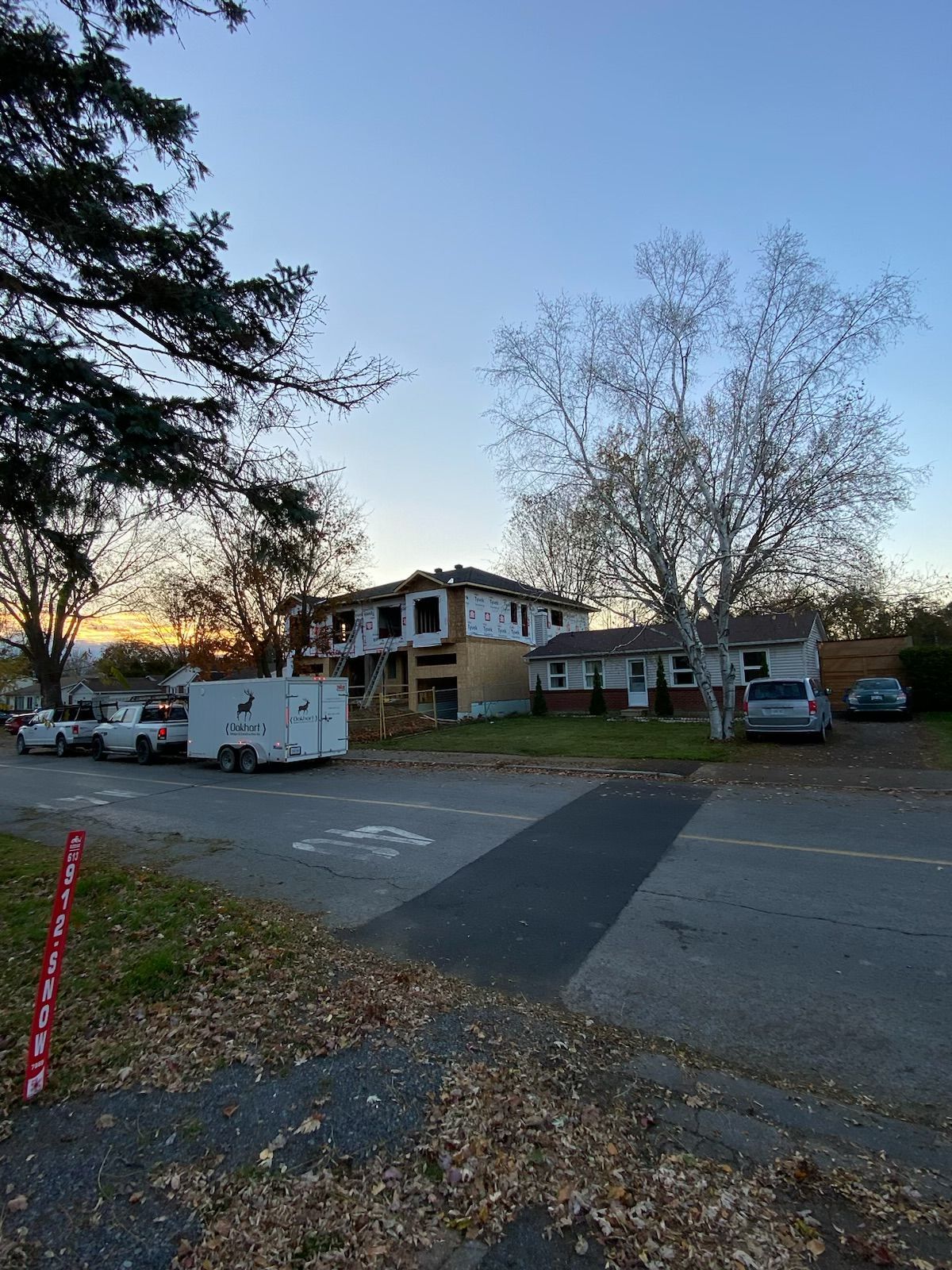 Residential street at dusk with parked vehicles and a two-story house behind leafless trees.