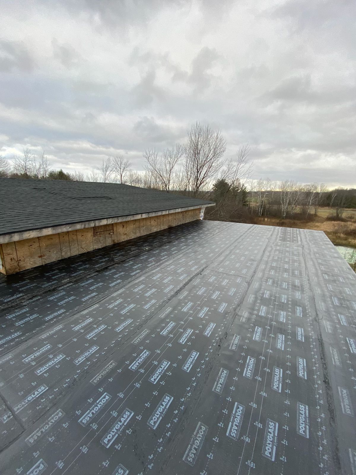 Roof underlayment on a sloped roof beside shingles, with cloudy sky and trees in the background