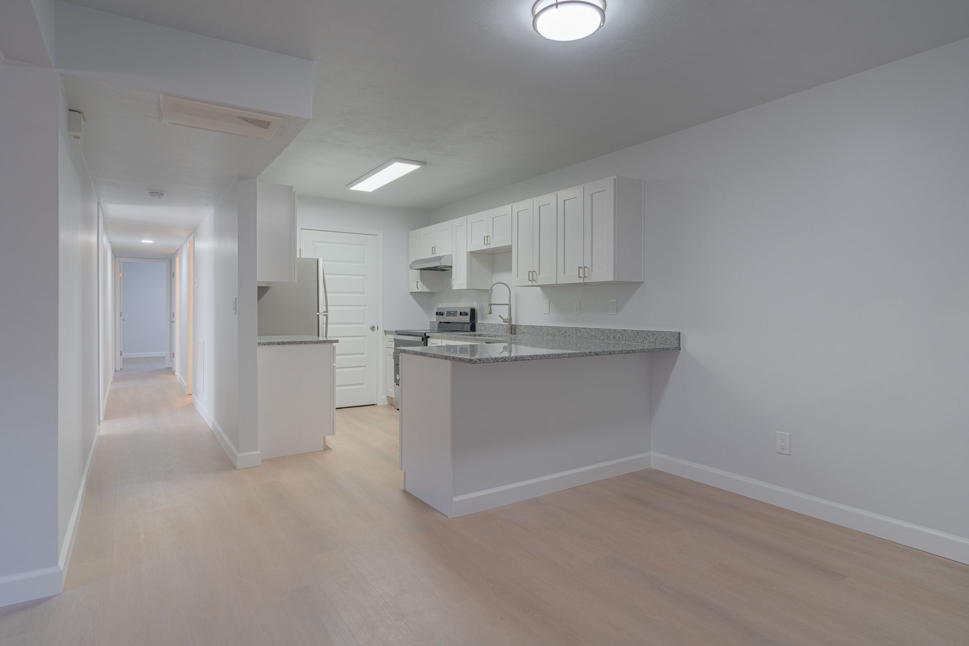 Bright empty kitchen with white cabinets, gray countertops, and light wood flooring in a hallway view