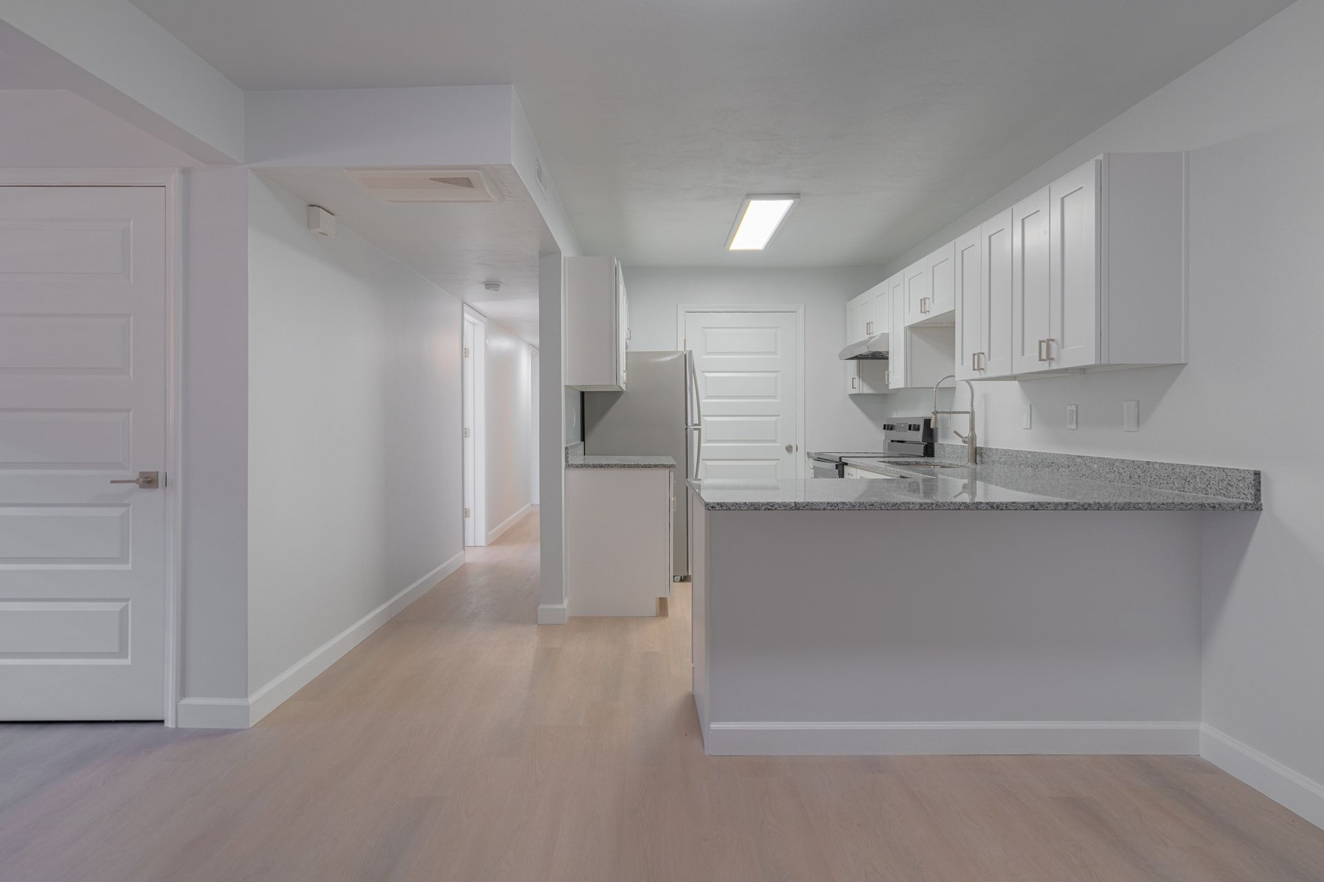 Empty modern kitchen with white cabinets, gray countertops, and light wood flooring, viewed from the entryway