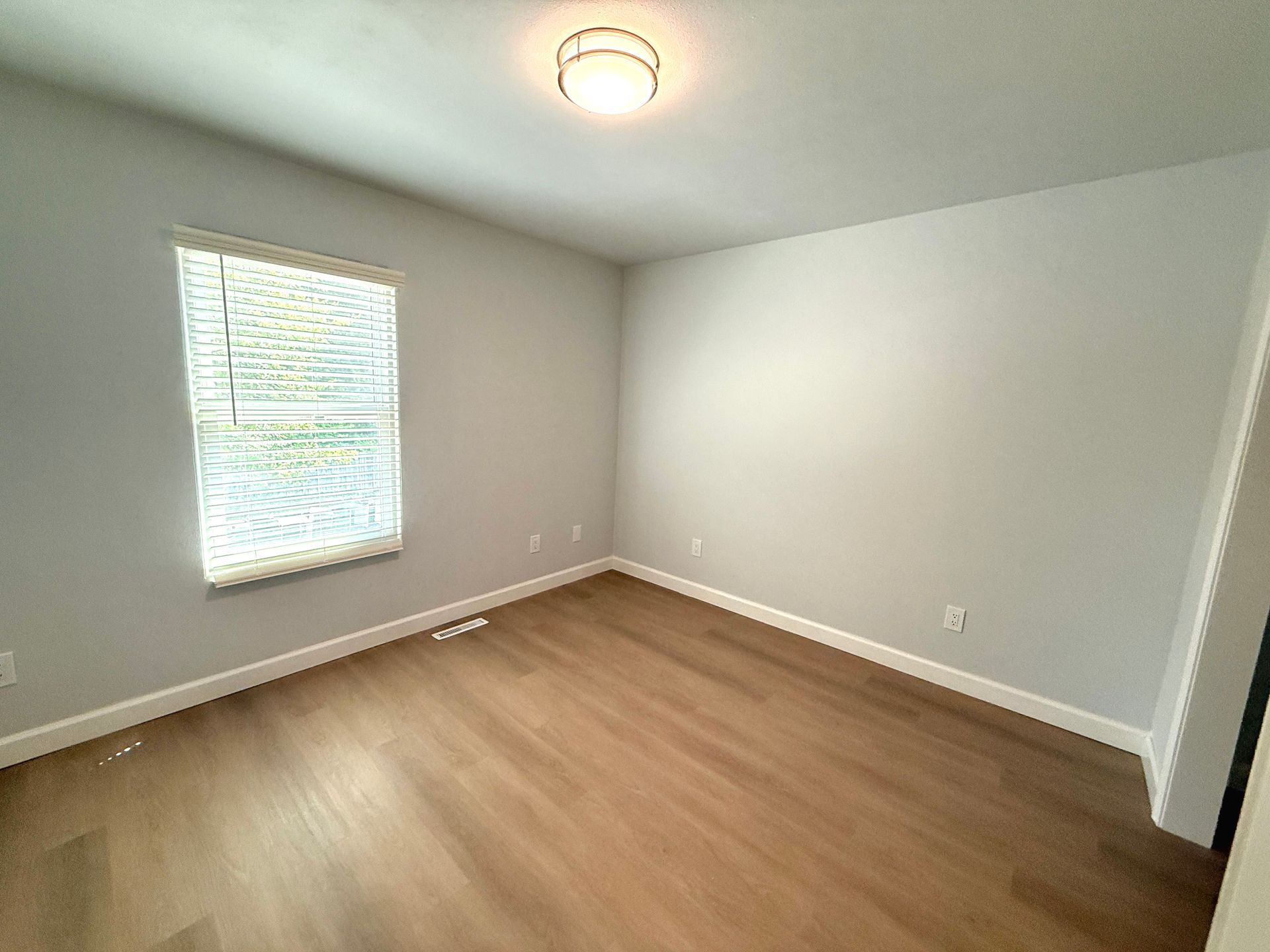Empty beige bedroom with wood floors, white walls, a window, and a ceiling light.