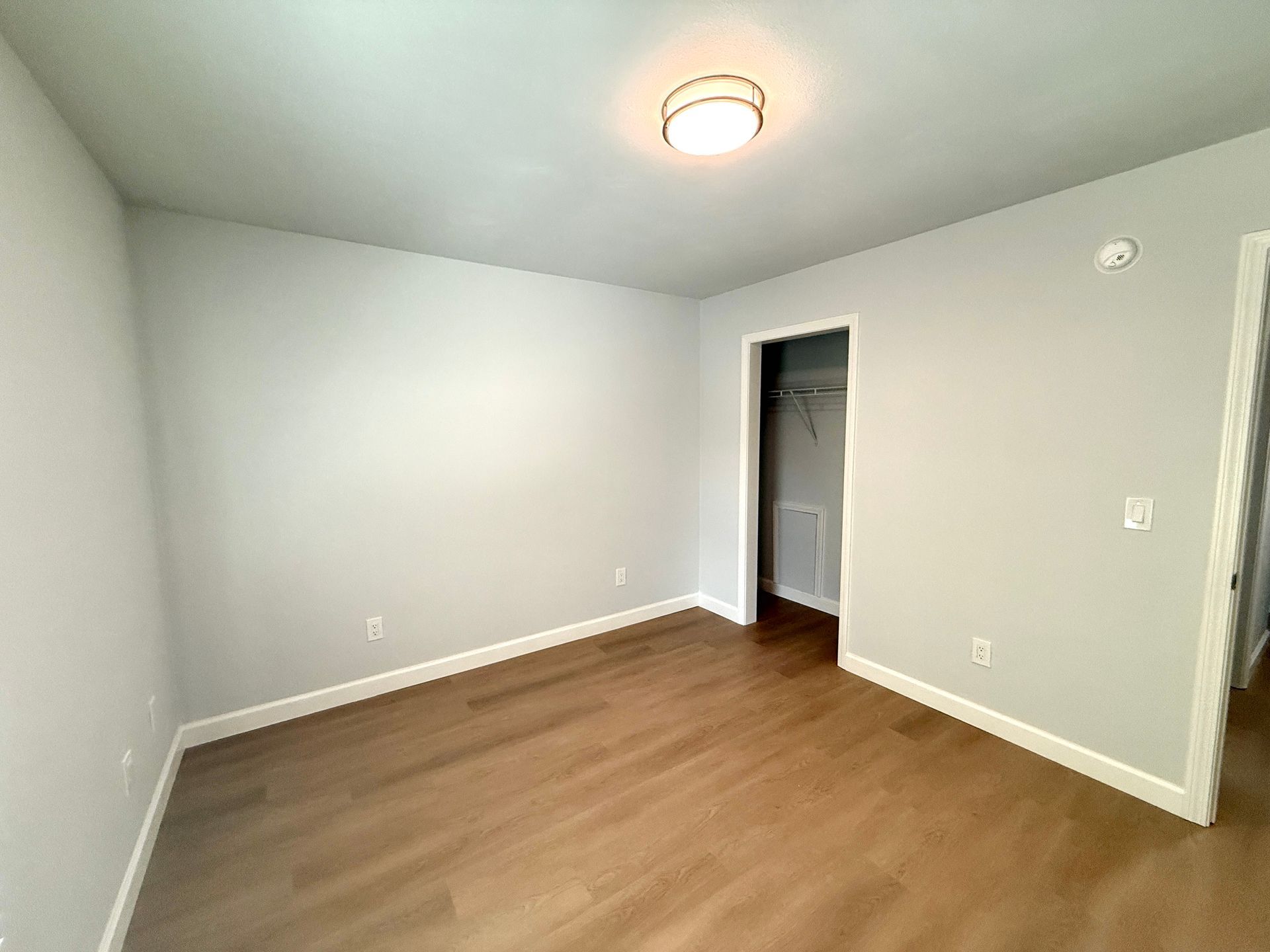 Empty gray bedroom with hardwood floors, white trim, and a closet doorway under a ceiling light.