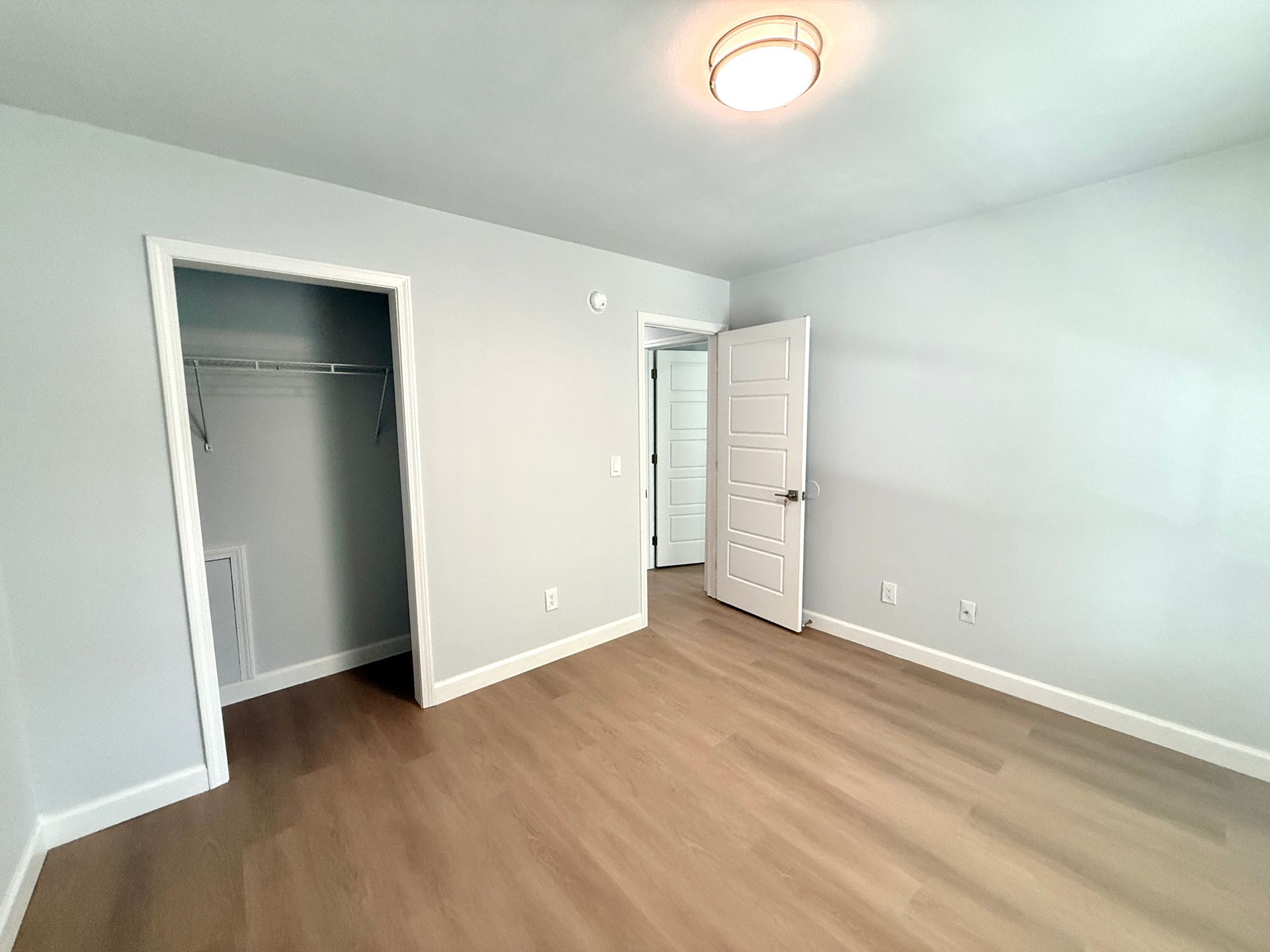 Empty bedroom with light gray walls, wood floors, open closet, and an open door to a hallway.