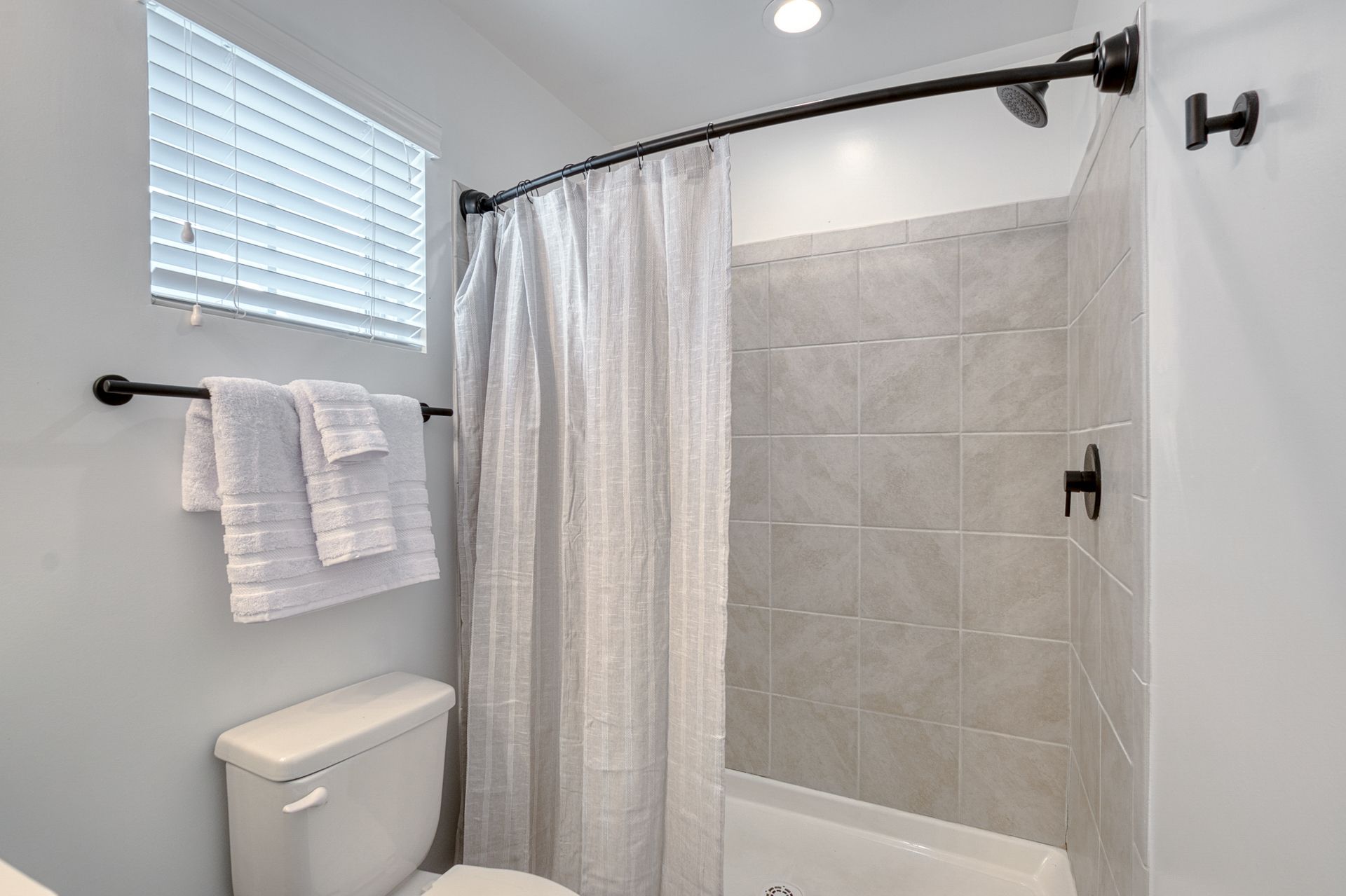 Small bright bathroom with walk-in shower, white curtain, towel rack, and window blinds.