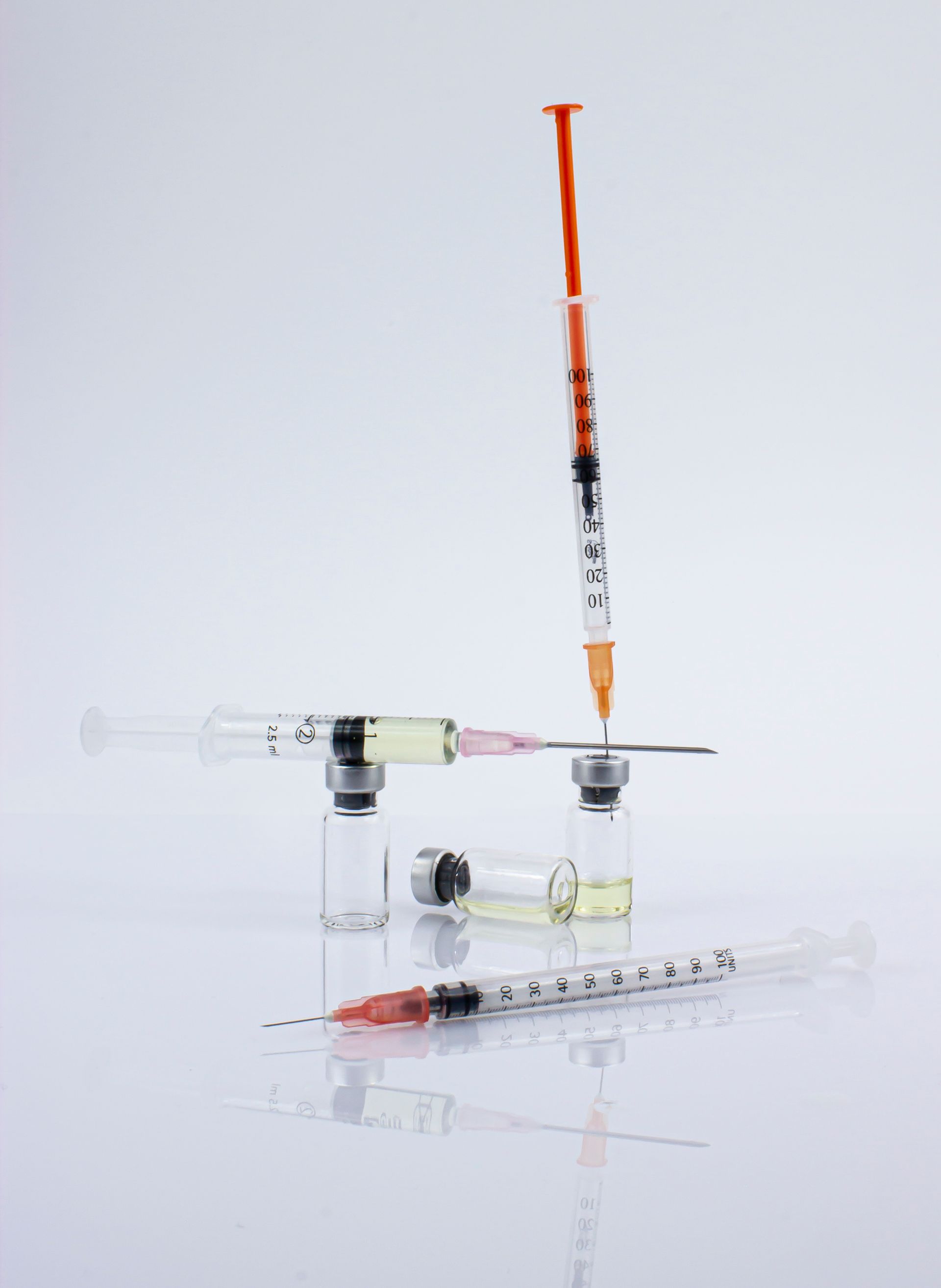 Two syringes and a small glass vial on a white surface, with one syringe standing upright and filled with orange liquid