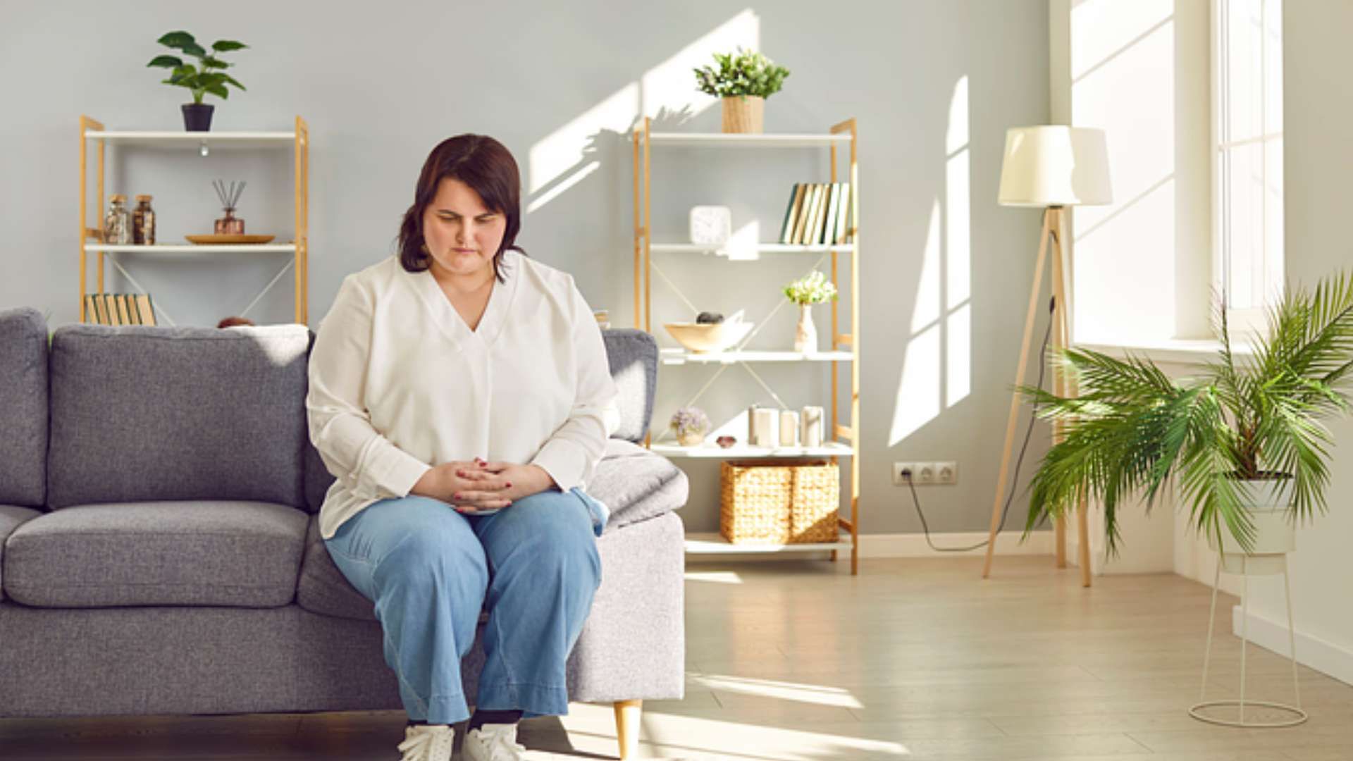 Woman sitting on a gray sofa in a bright living room, looking down thoughtfully.