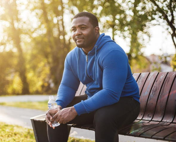 Man sitting on a bench outdoors in a blue hoodie, holding a water bottle in warm sunlight