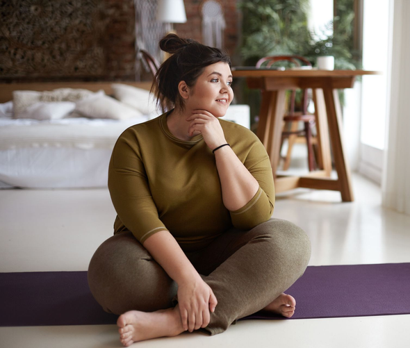 Woman seated cross-legged on a yoga mat in a bright room, looking off to the side.
