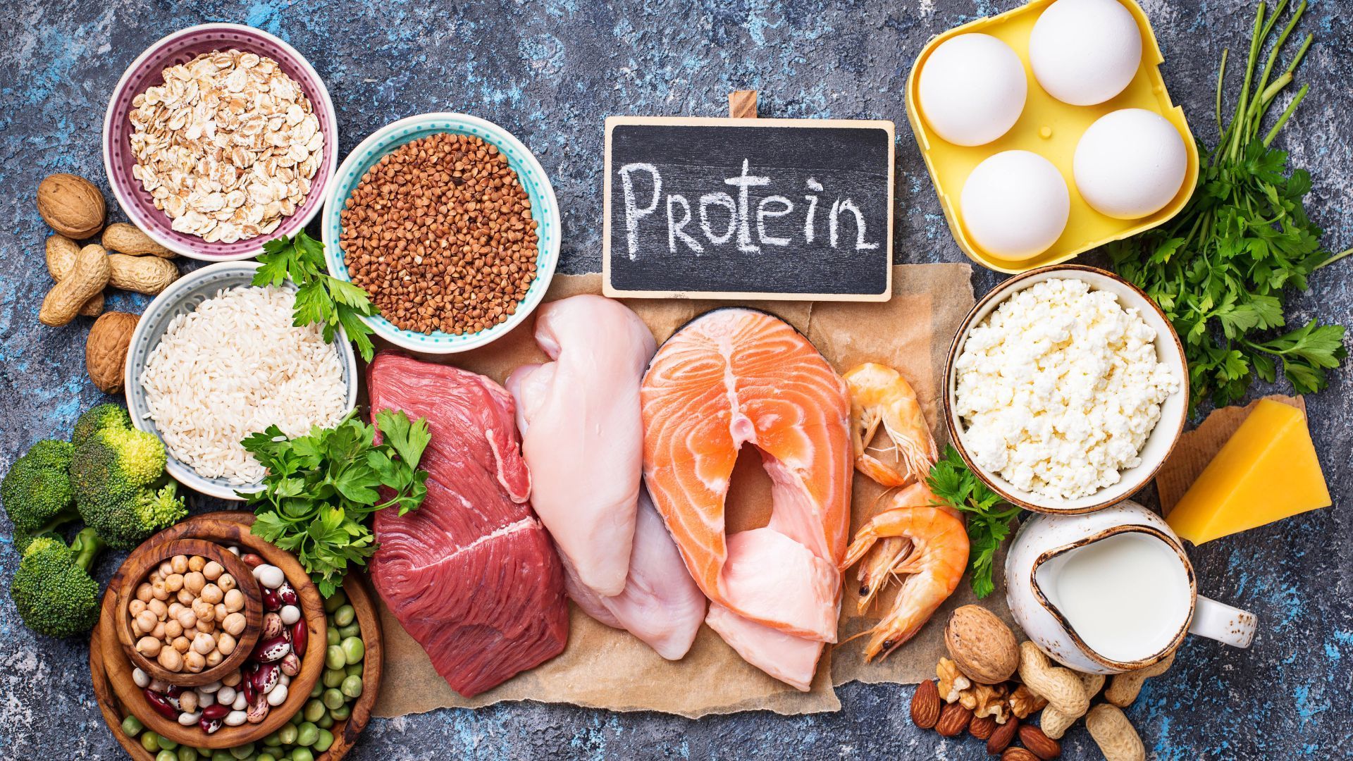 Assorted protein foods with chalkboard label, including eggs, meat, fish, cheese, beans, and nuts on a tabletop