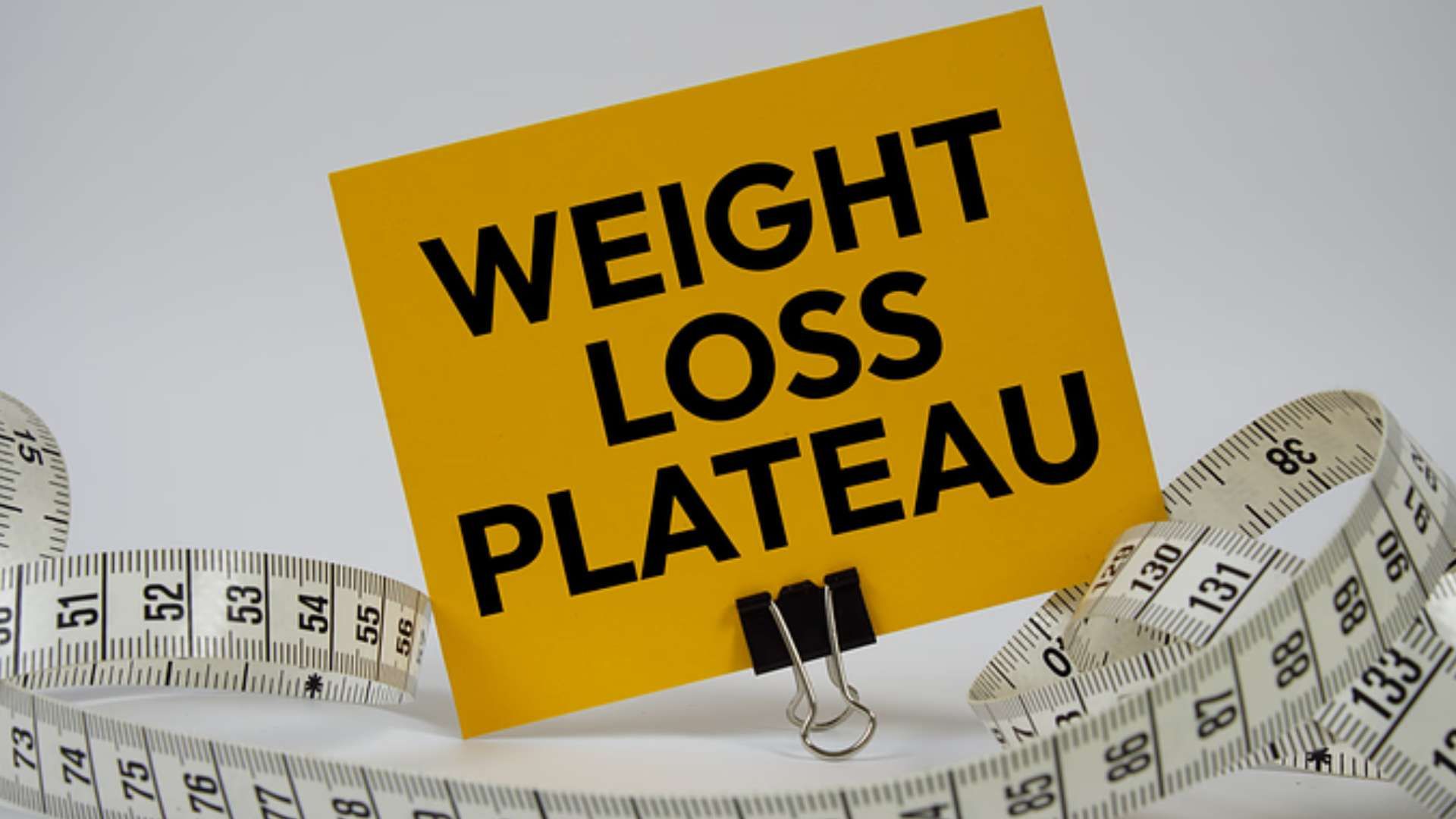 Yellow sign reading “WEIGHT LOSS PLATEAU” over a measuring tape background