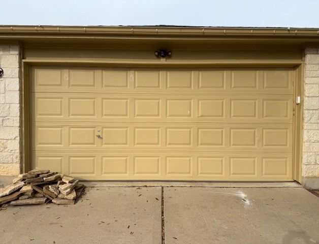 Beige garage door on a concrete driveway with stacked firewood on the left