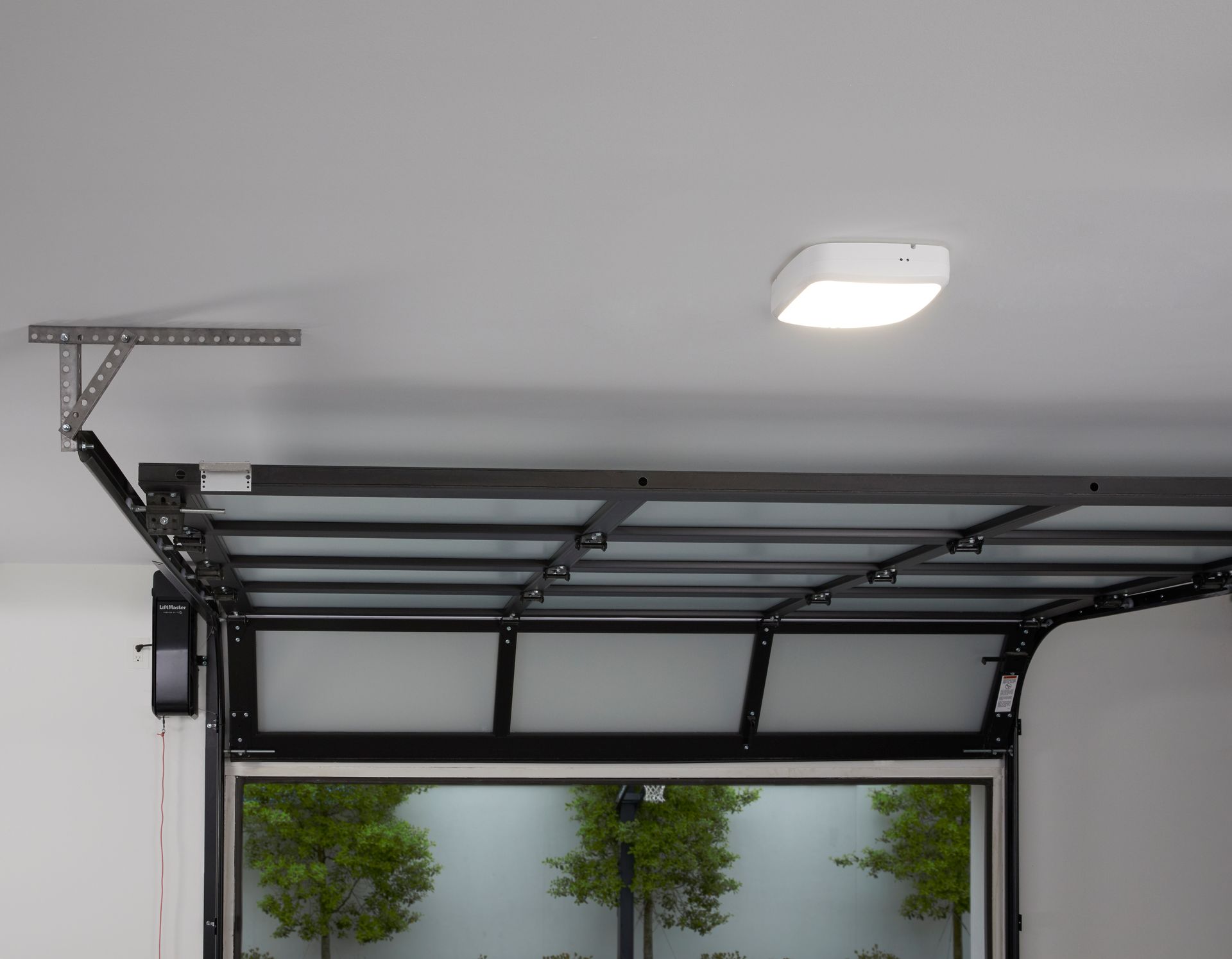 Open garage with dark metal door frame and white ceiling light above two potted trees outside.