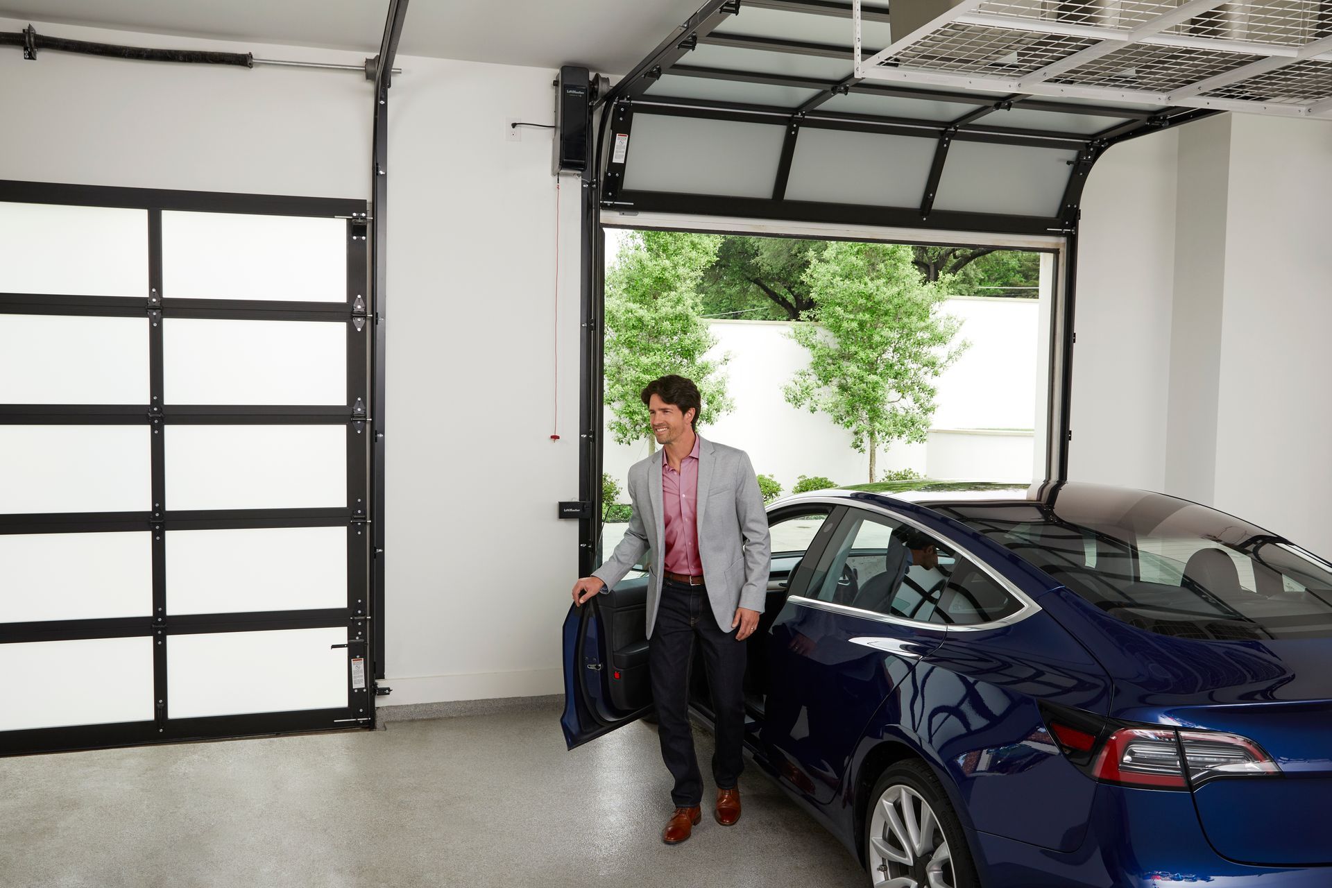 Man standing beside a blue car in an open garage, holding a bag.