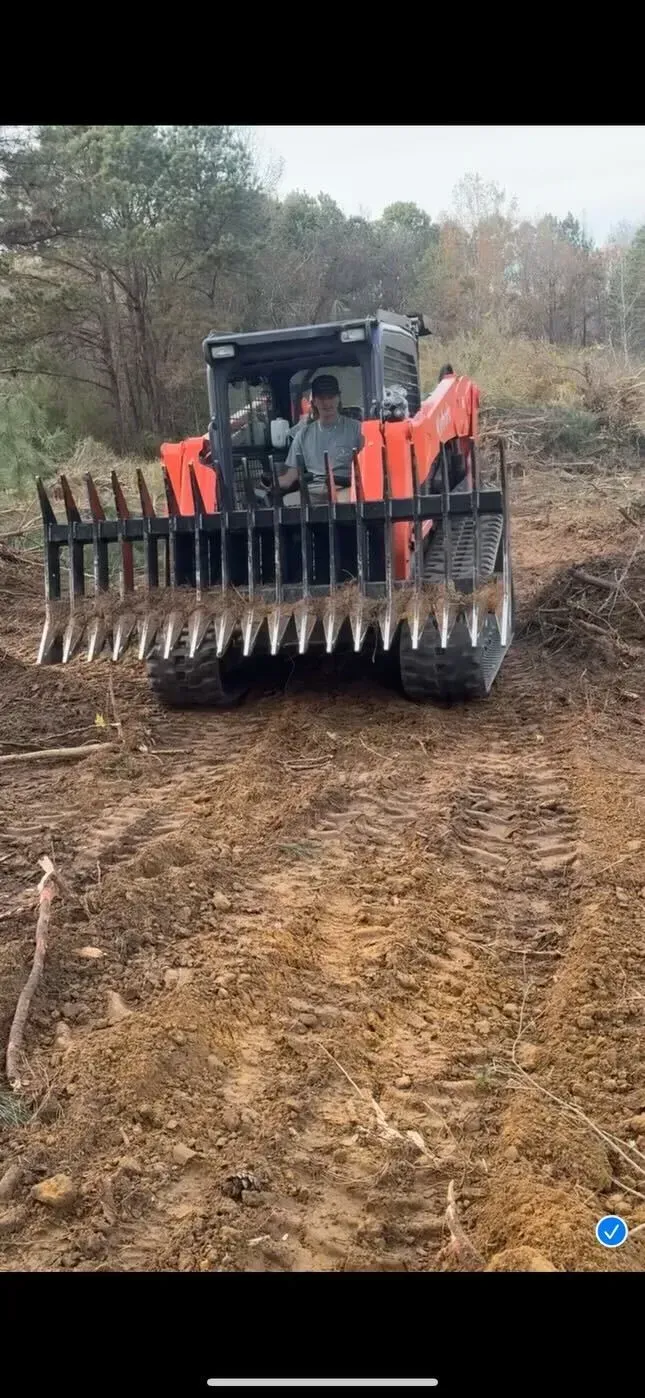 Orange tractor-mounted logging rake on muddy dirt road in a wooded area