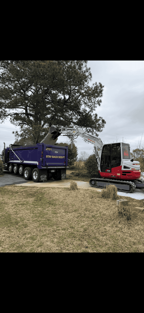 Purple dump truck and red excavator parked on a dirt lot near a tree under cloudy sky