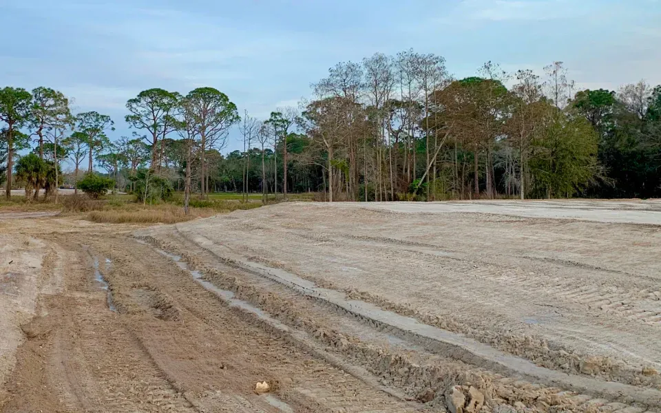 Cleared dirt lot with tire tracks beside a line of trees under a cloudy sky