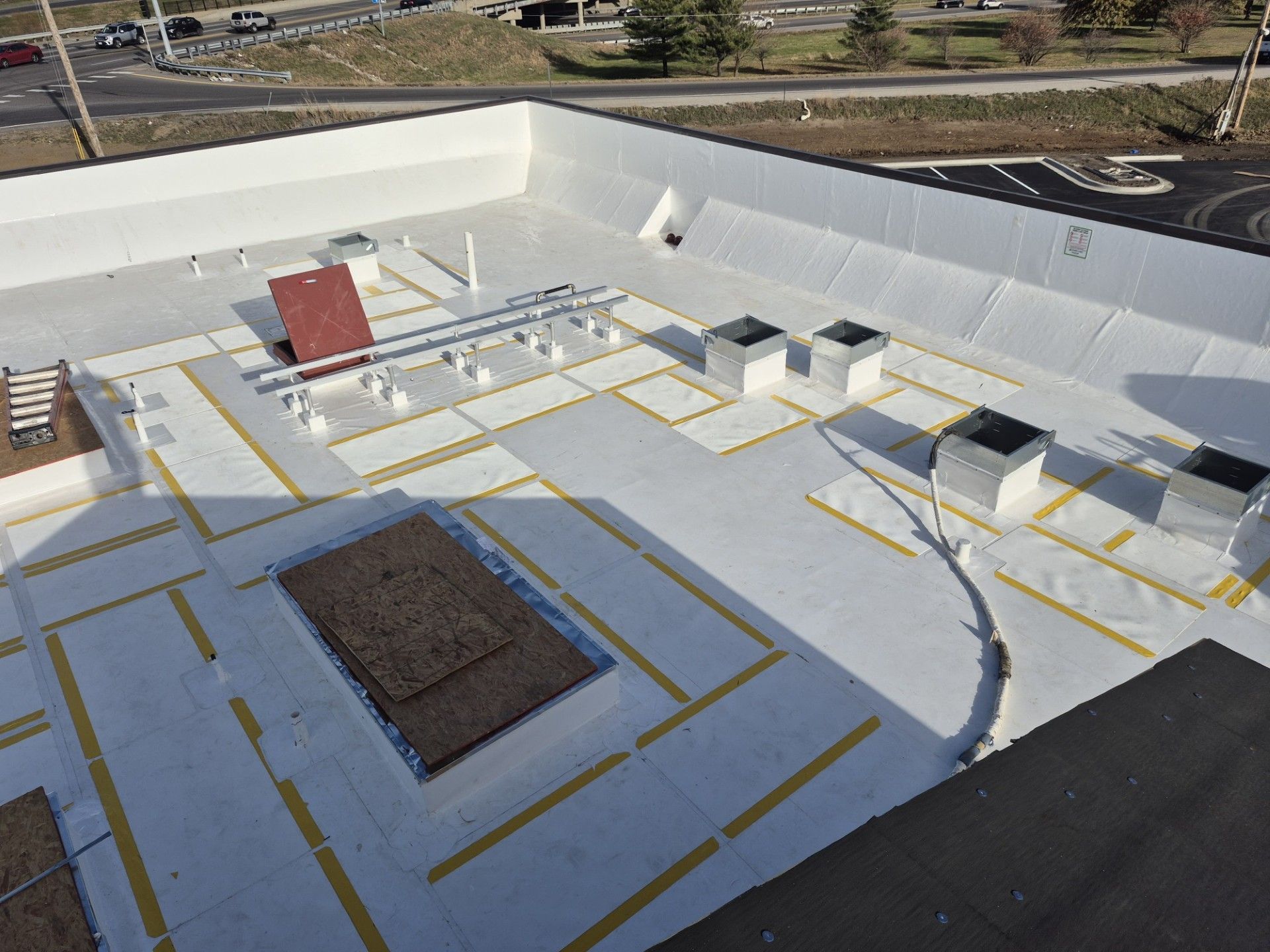 Rooftop construction area with white waterproofing, yellow tape lines, vents, and a red equipment box
