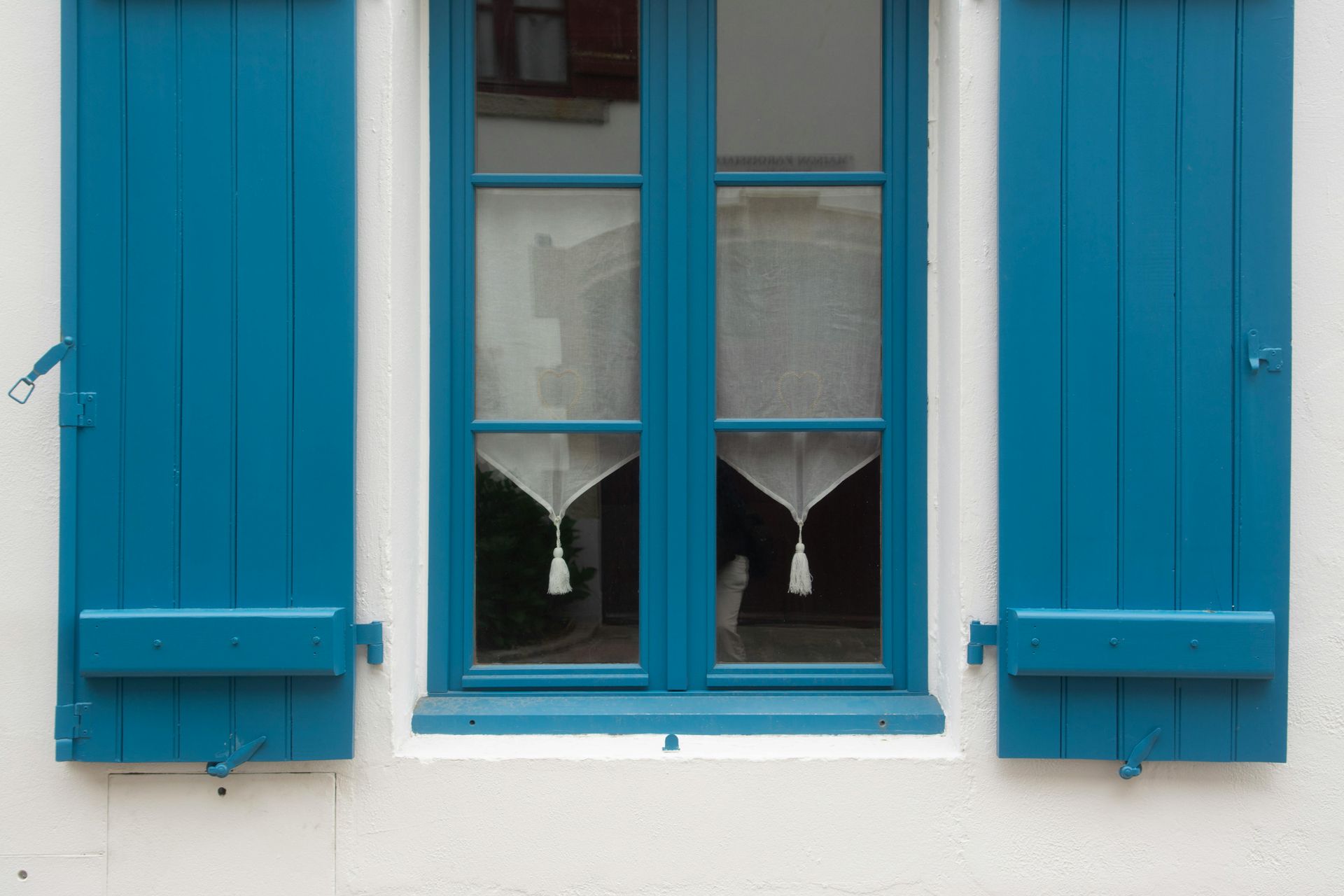 Blue window shutters flank a blue-framed window with white curtains.