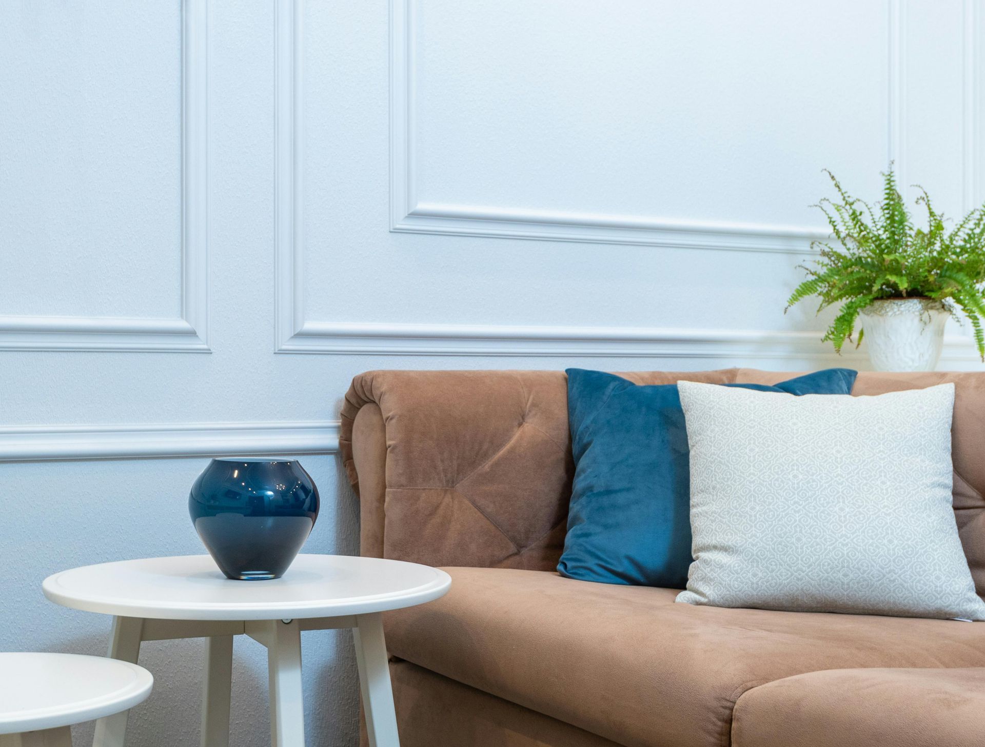 A brown sofa with blue and white pillows next to a white side table with a dark blue vase, and a fern.