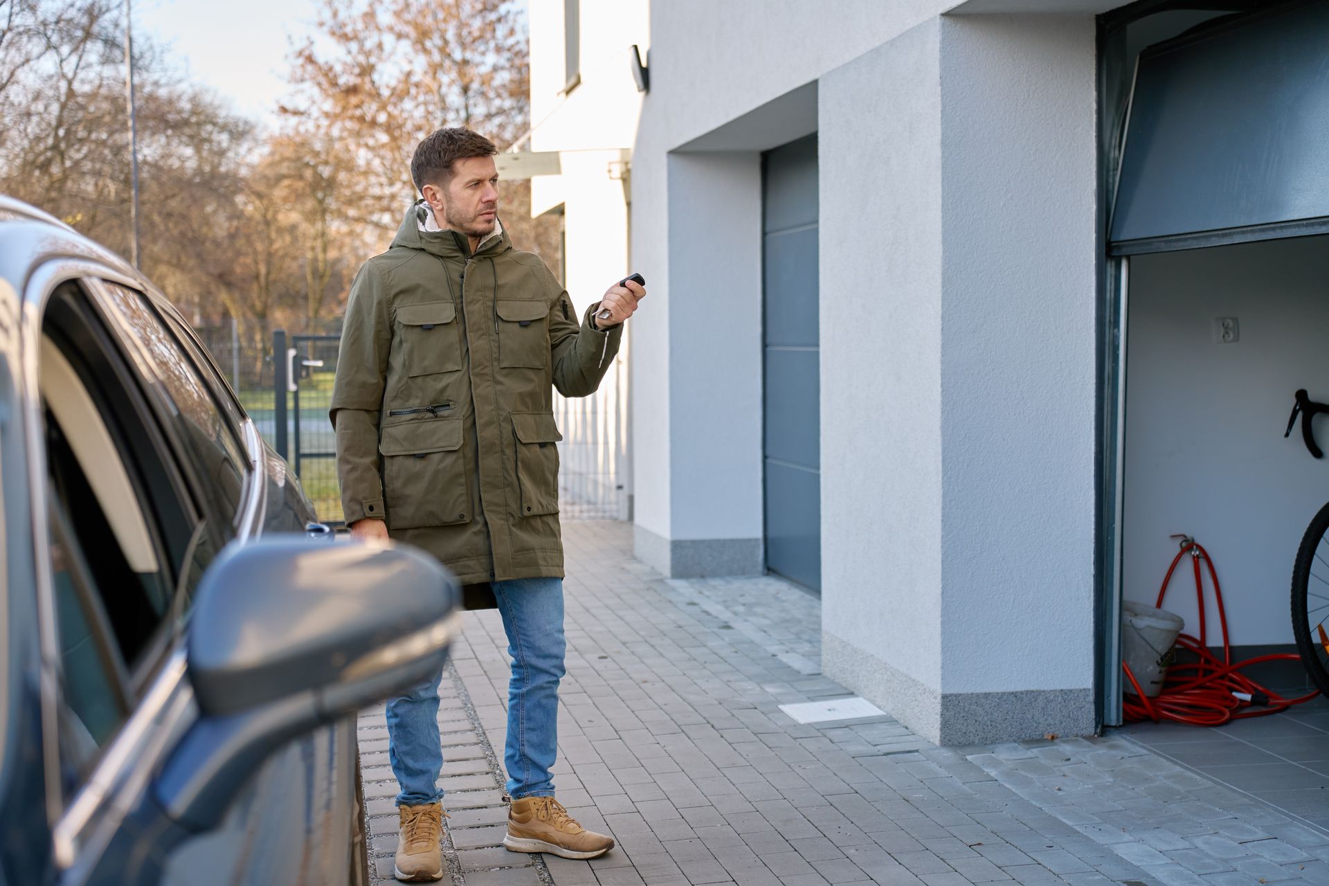 A man is standing outside a house near a parked car, using a remote control to open a garage door.