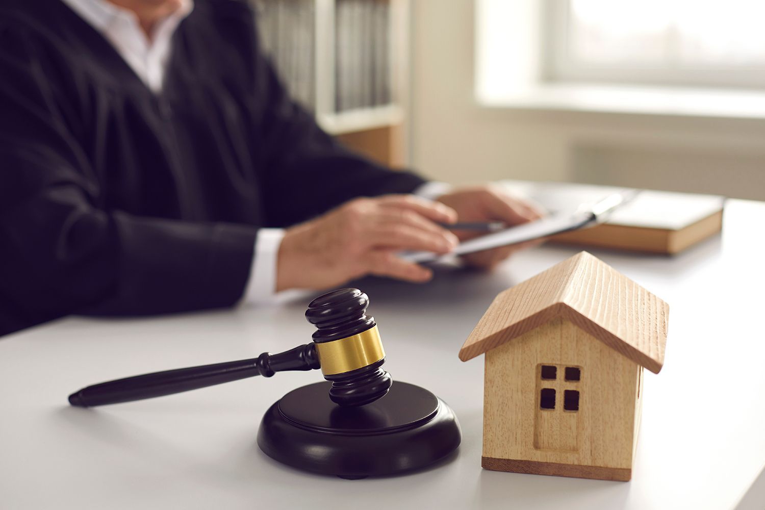 Judge’s gavel beside a small house model on a desk with a person writing in the background Judge’s gavel beside a small house model on a desk with a person writing in the background