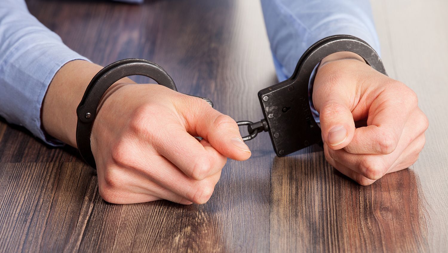 Hands in handcuffs resting on a wooden table, with a person wearing a light blue shirt. Hands in handcuffs resting on a wooden table, with a person wearing a light blue shirt.