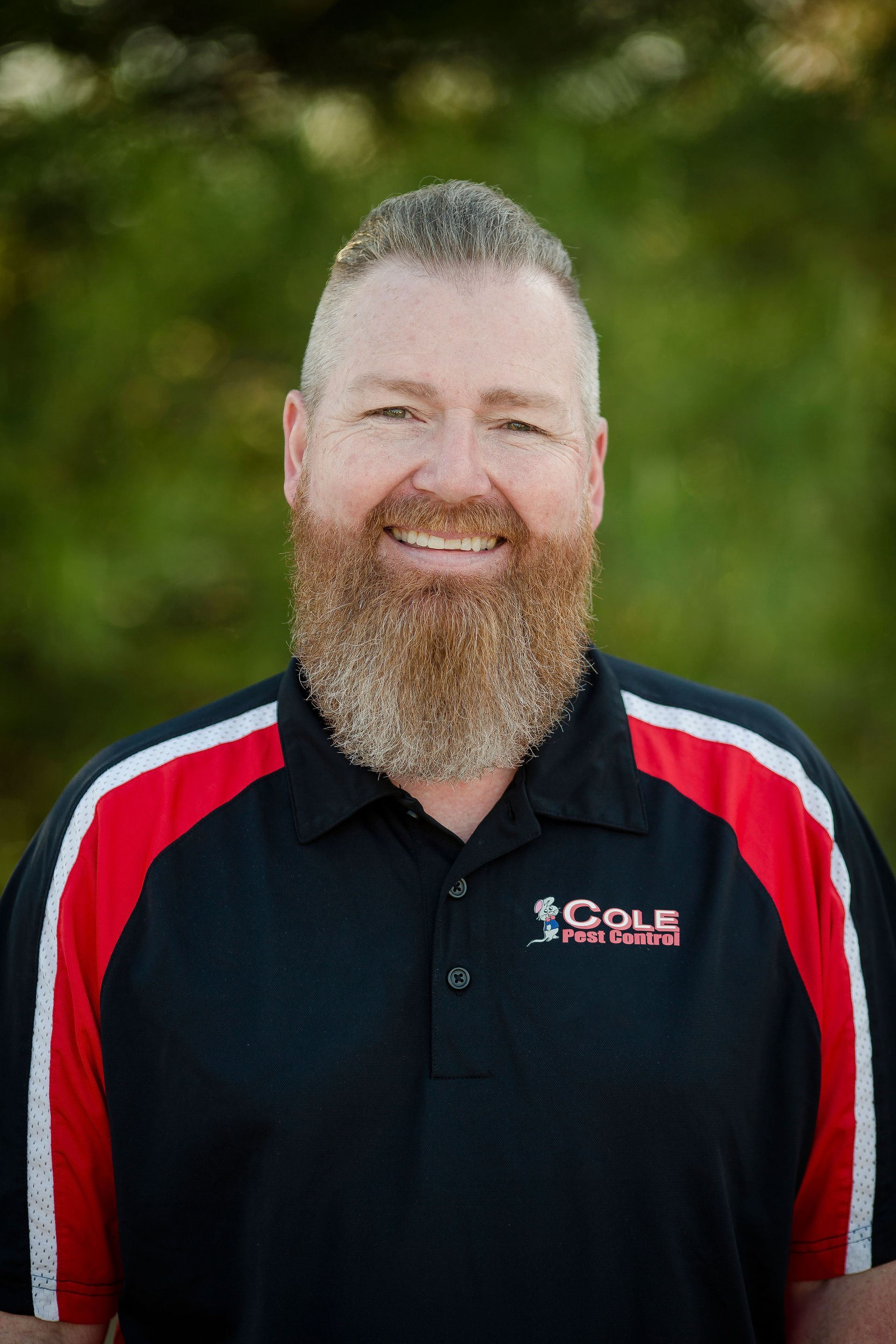 Smiling man in black Cole shirt with red and white stripes, standing outdoors against a blurred green background