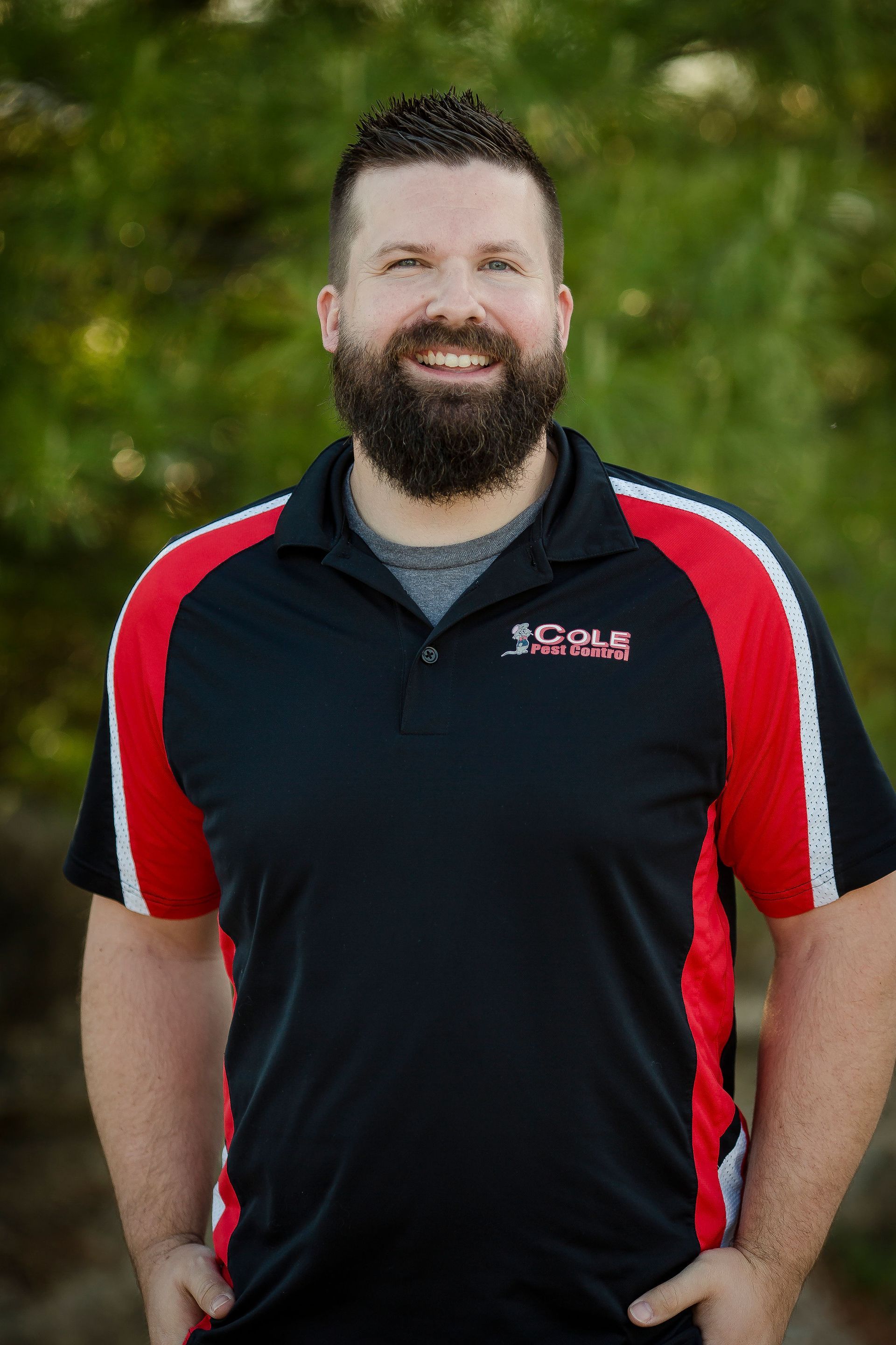 Smiling man in a black polo with red stripes standing outdoors against a green background