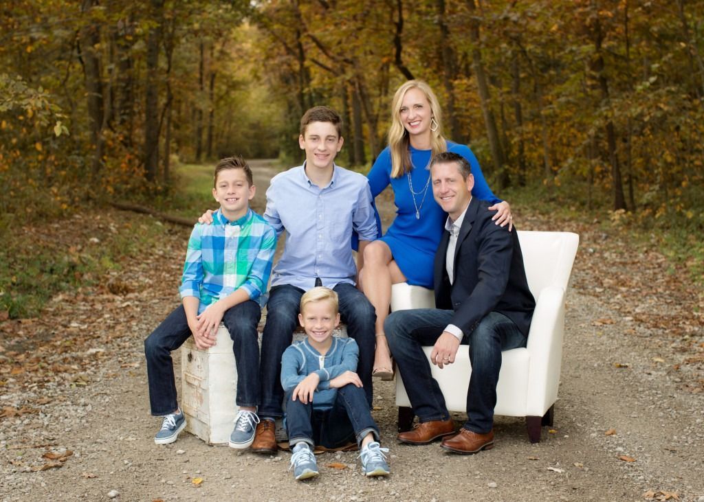 Family portrait of five on white couches outdoors in autumn woods, with relaxed smiles.