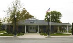 Small brick building with a covered porch, trees, and an American flag by the sidewalk