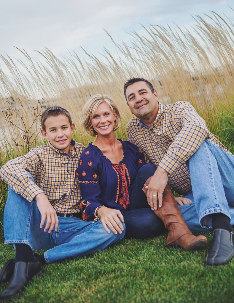 Three people sitting on grass in front of tall ornamental grasses, smiling at the camera.