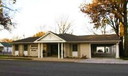 Single-story beige building with a covered front porch and carport, set beside a road with trees nearby