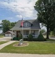 Small gray house with a porch, American flag, and front yard garden under a cloudy sky
