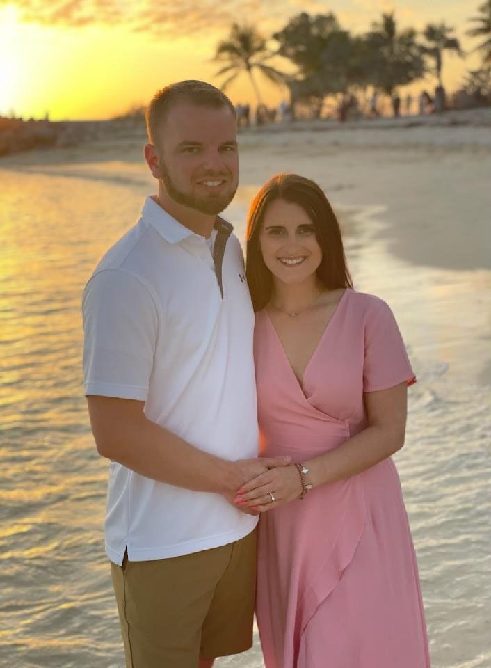 Couple standing on a beach at sunset, smiling and holding hands.