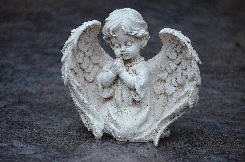 Small white angel figurine with folded hands and large wings on a dark fabric background