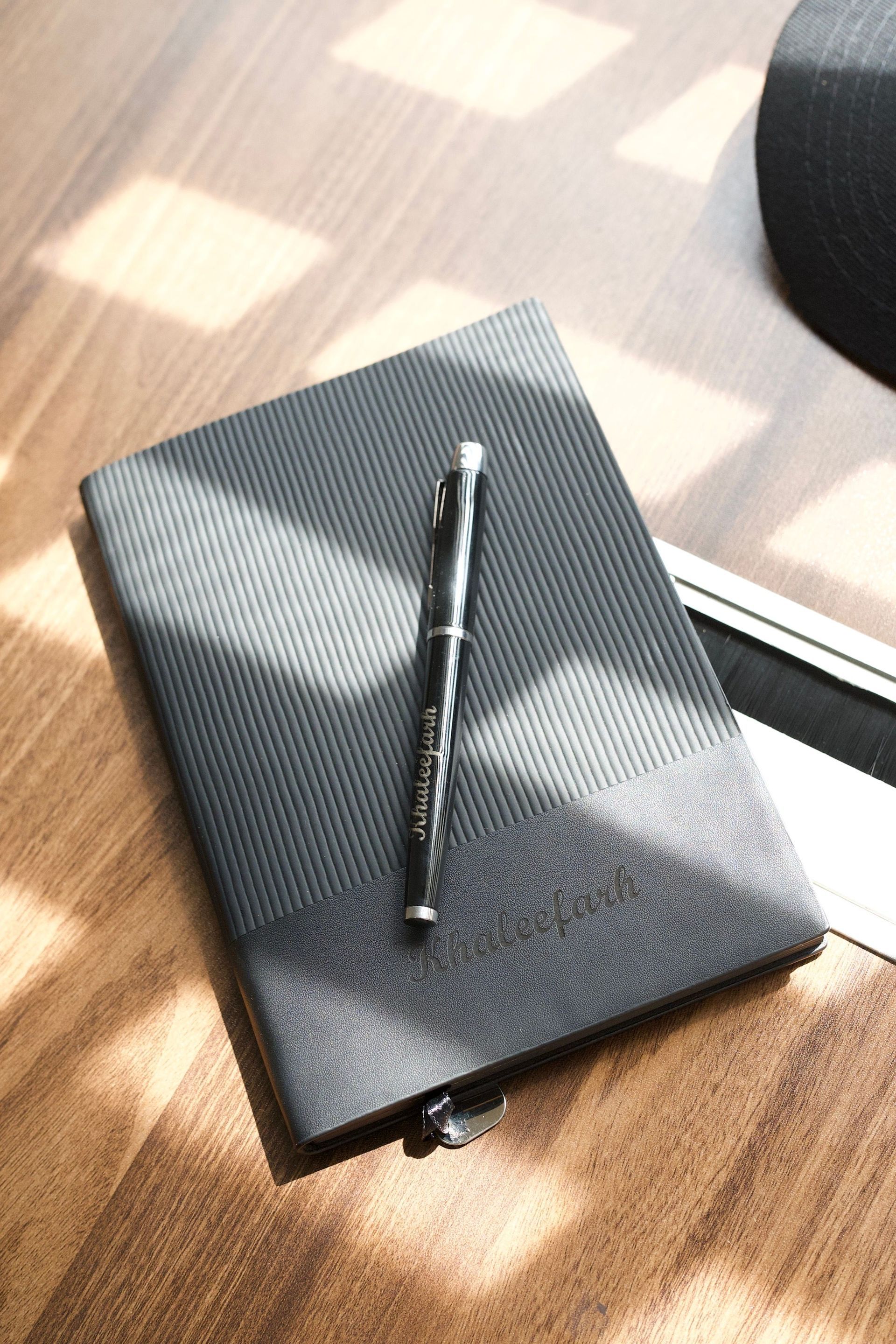 Black notebook on a wooden table with a silver pen resting on top.