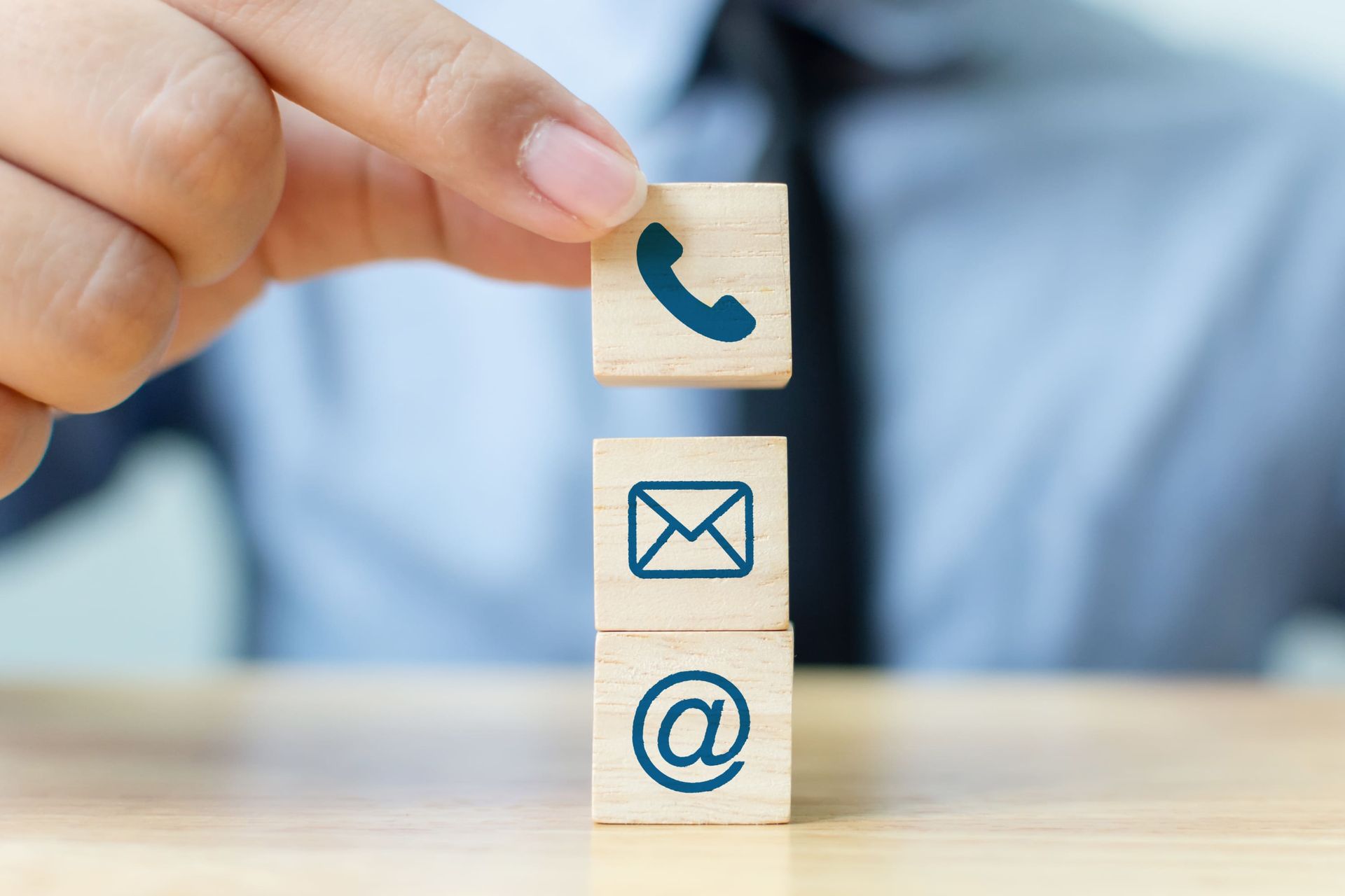 A hand placing a wooden block with a phone icon on top of two others displaying email and at-symbol icons.