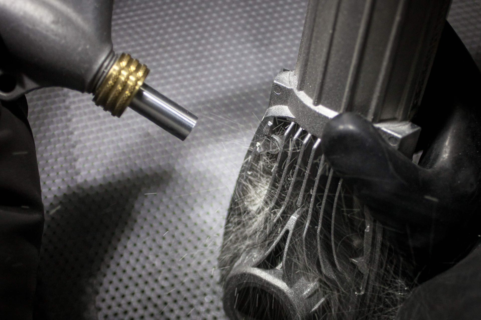 A gloved hand holds a metal component as a nozzle cleans it with pressurized air in an industrial setting.