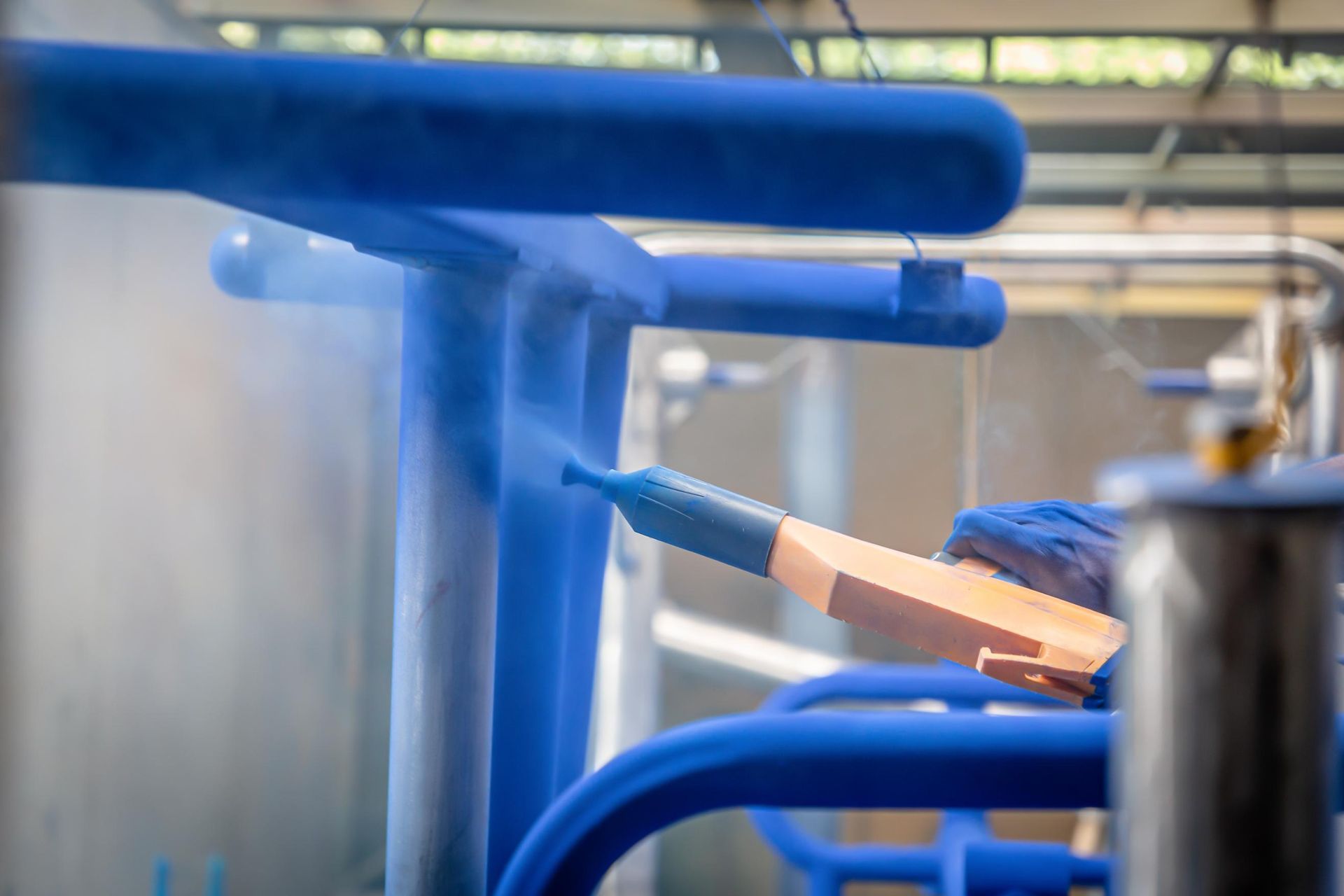 A gloved hand uses a spray gun to apply blue powder coating to a metal frame in an industrial workshop.
