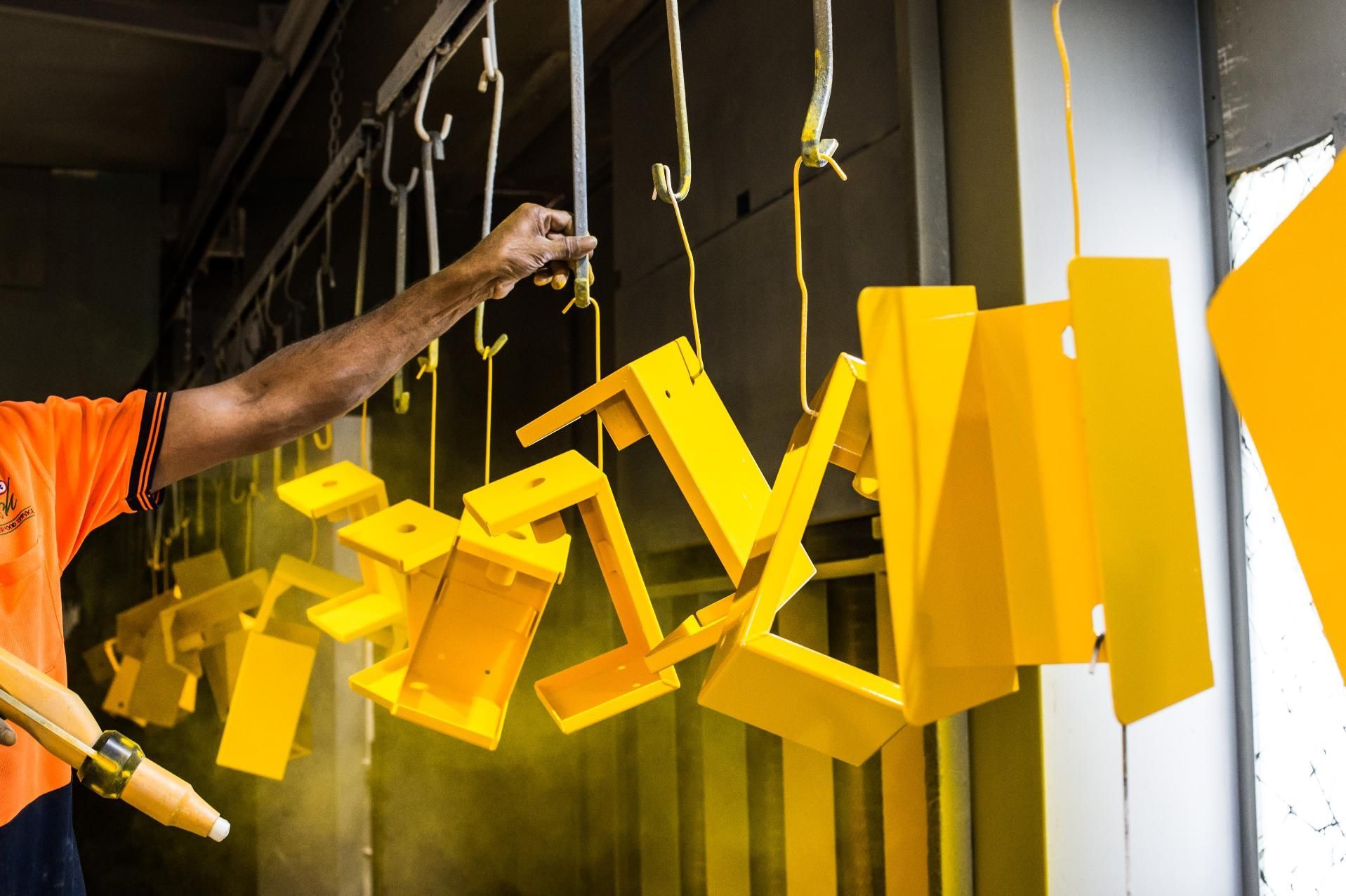 A worker uses a spray gun to apply yellow powder coating to metal parts hanging on a conveyor line.